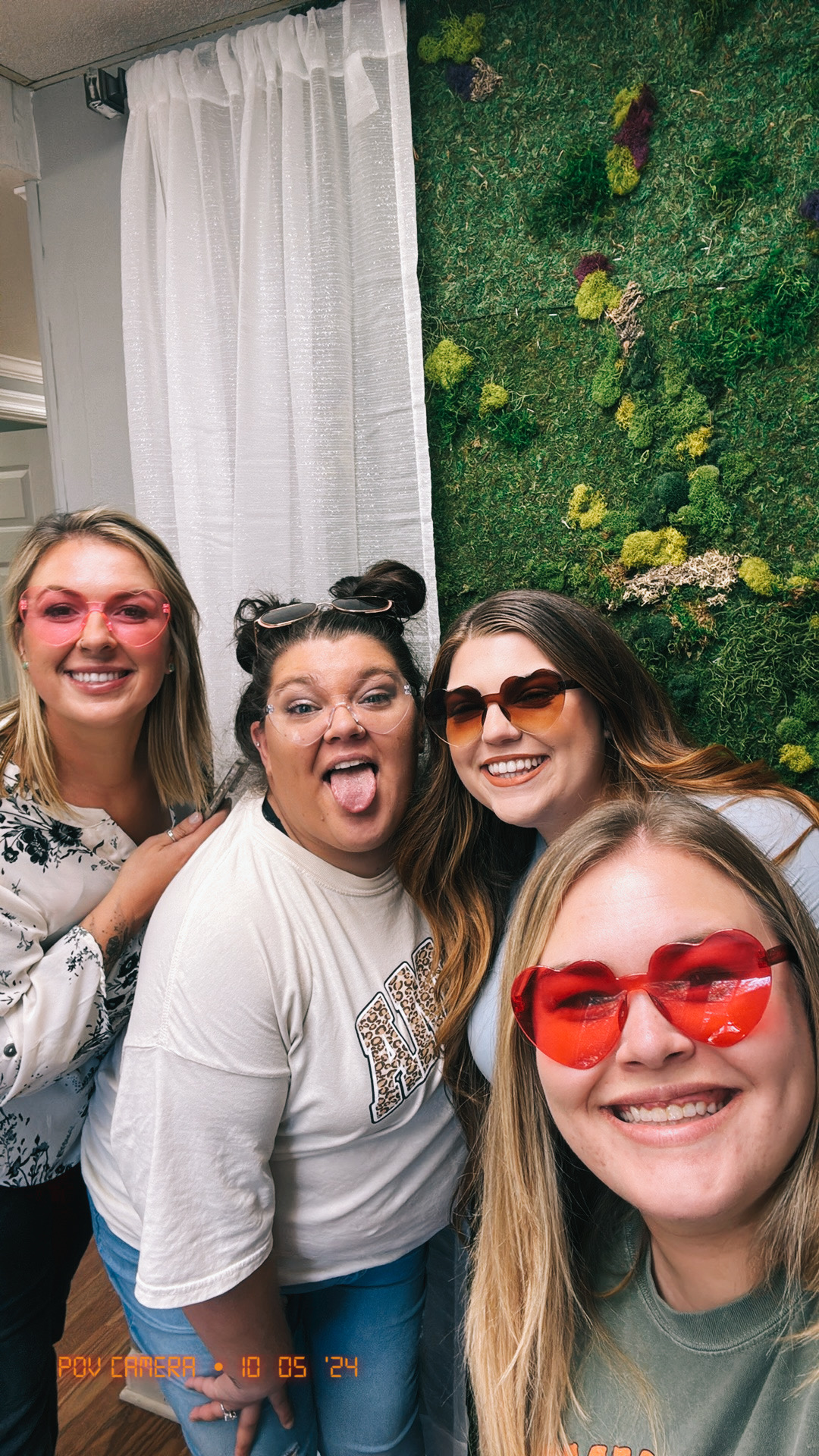 Four women smiling and posing for a photo indoors, one wearing red sunglasses, with a green wall and white curtain behind them.