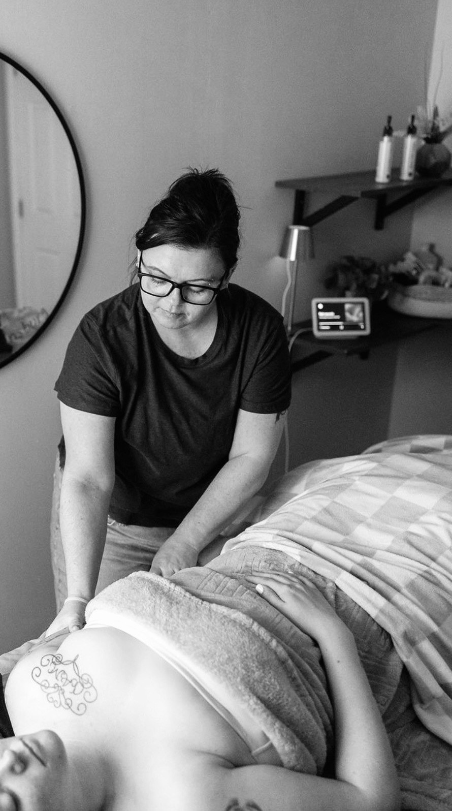 Woman with glasses and dark hair in a black shirt leans over a person lying on a bed, in a room with a mirror and shelf.