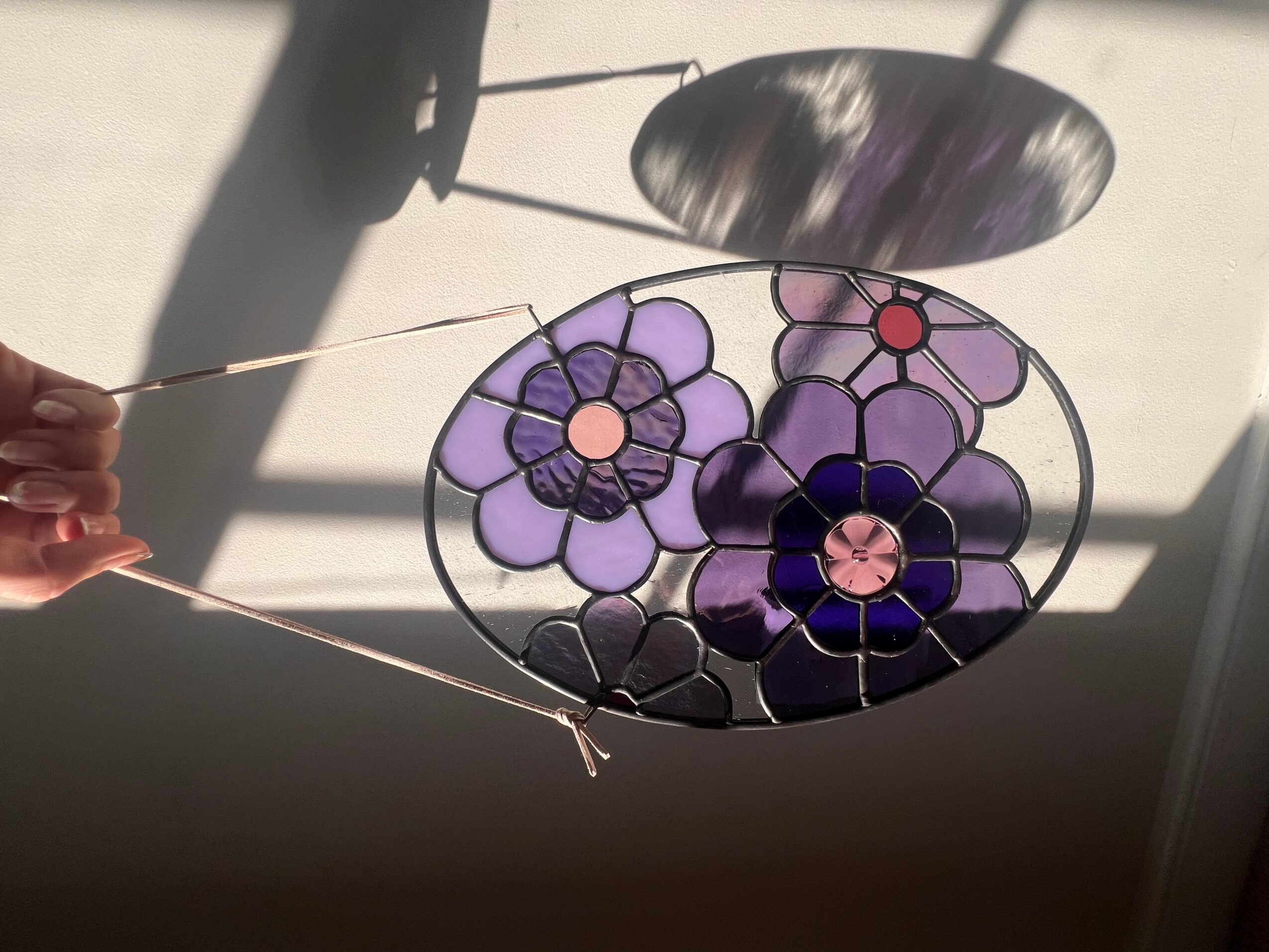 Colorful stained glass flower design with purple, pink, and red elements, hanging in front of a window with sunlight and shadows.