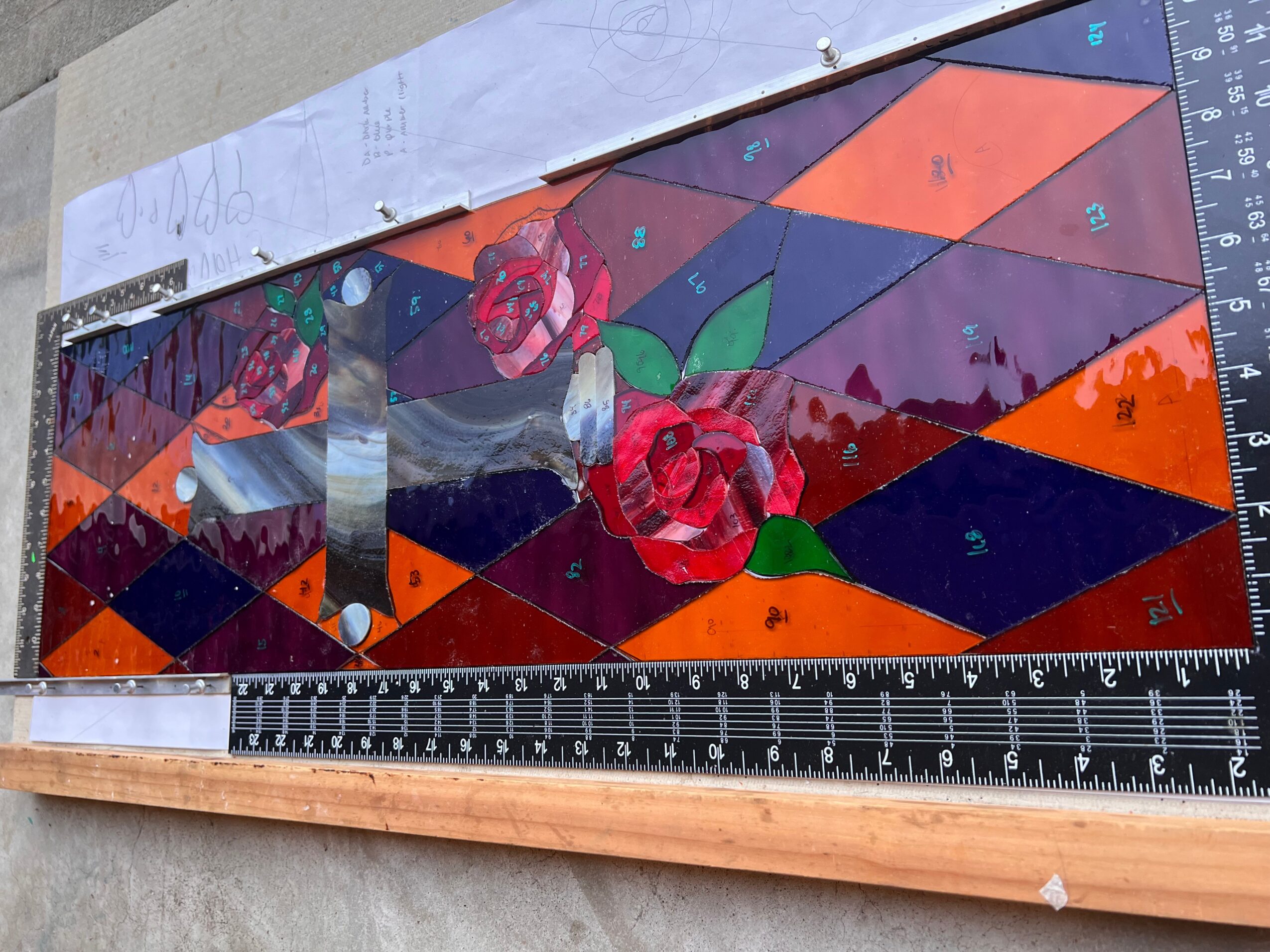 Colorful geometric stained glass artwork with roses, mountains, and abstract shapes, on a measuring tool background.