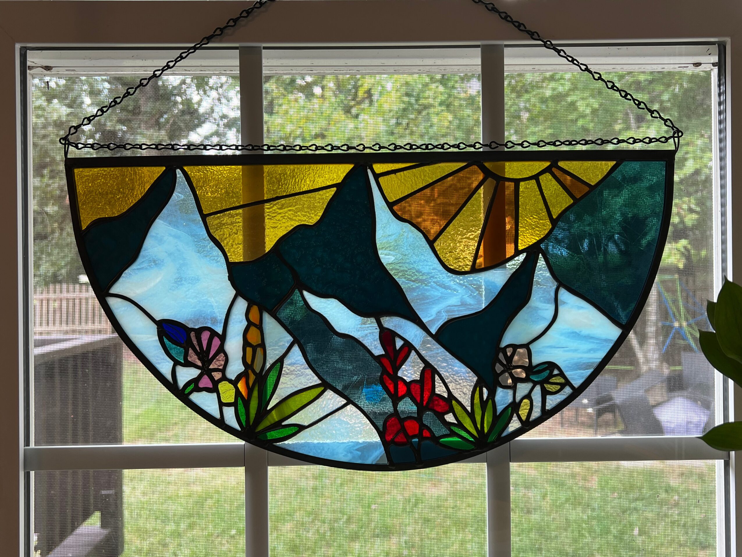 Stained glass window with mountains, sun, and flowers, hanging in front of a window with trees outside.