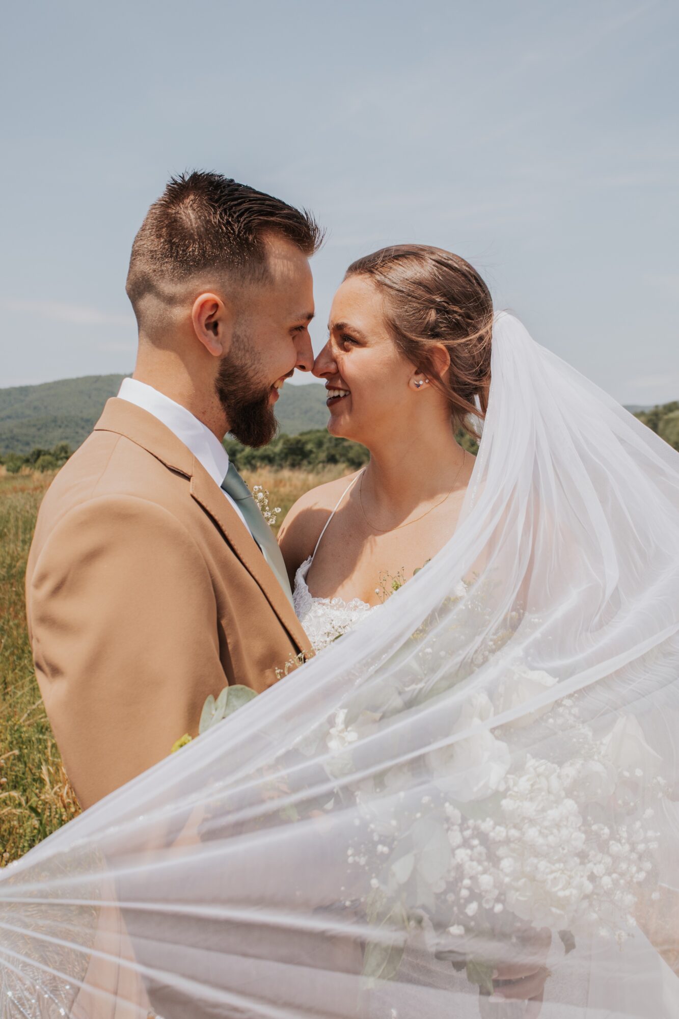 A bride and groom face each other outdoors, smiling, with a landscape and sky in the background.