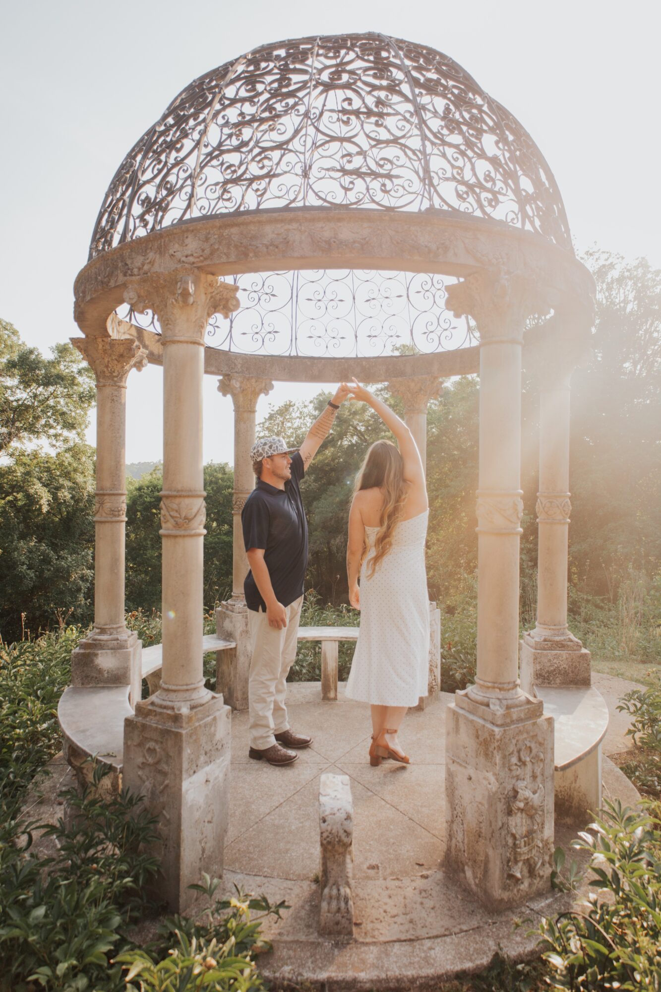 Two people stand under a decorative gazebo with a domed metal roof, holding hands, surrounded by greenery and sunlight.