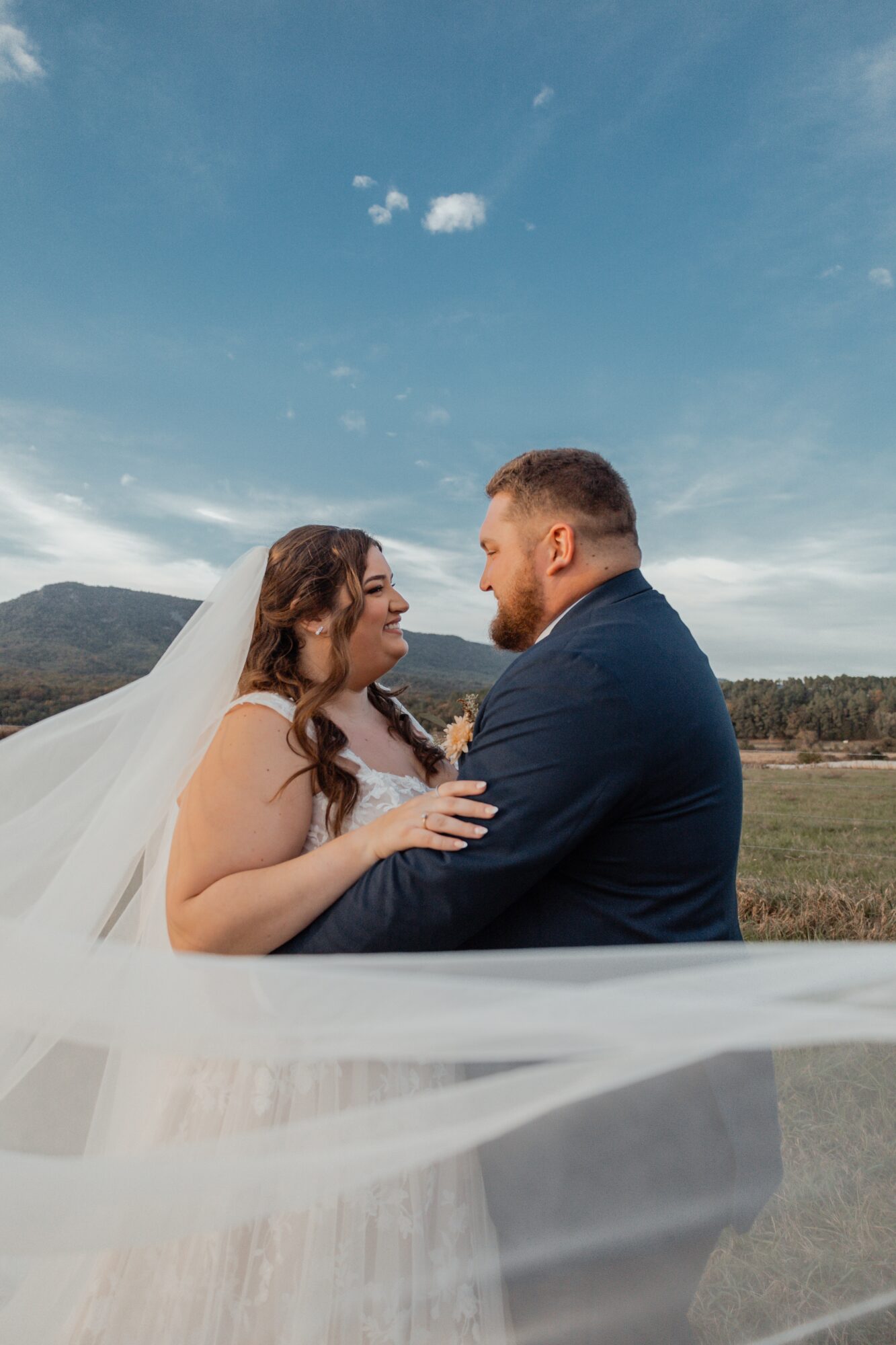 Bride and groom face each other outdoors under a blue sky with clouds, holding hands and smiling.