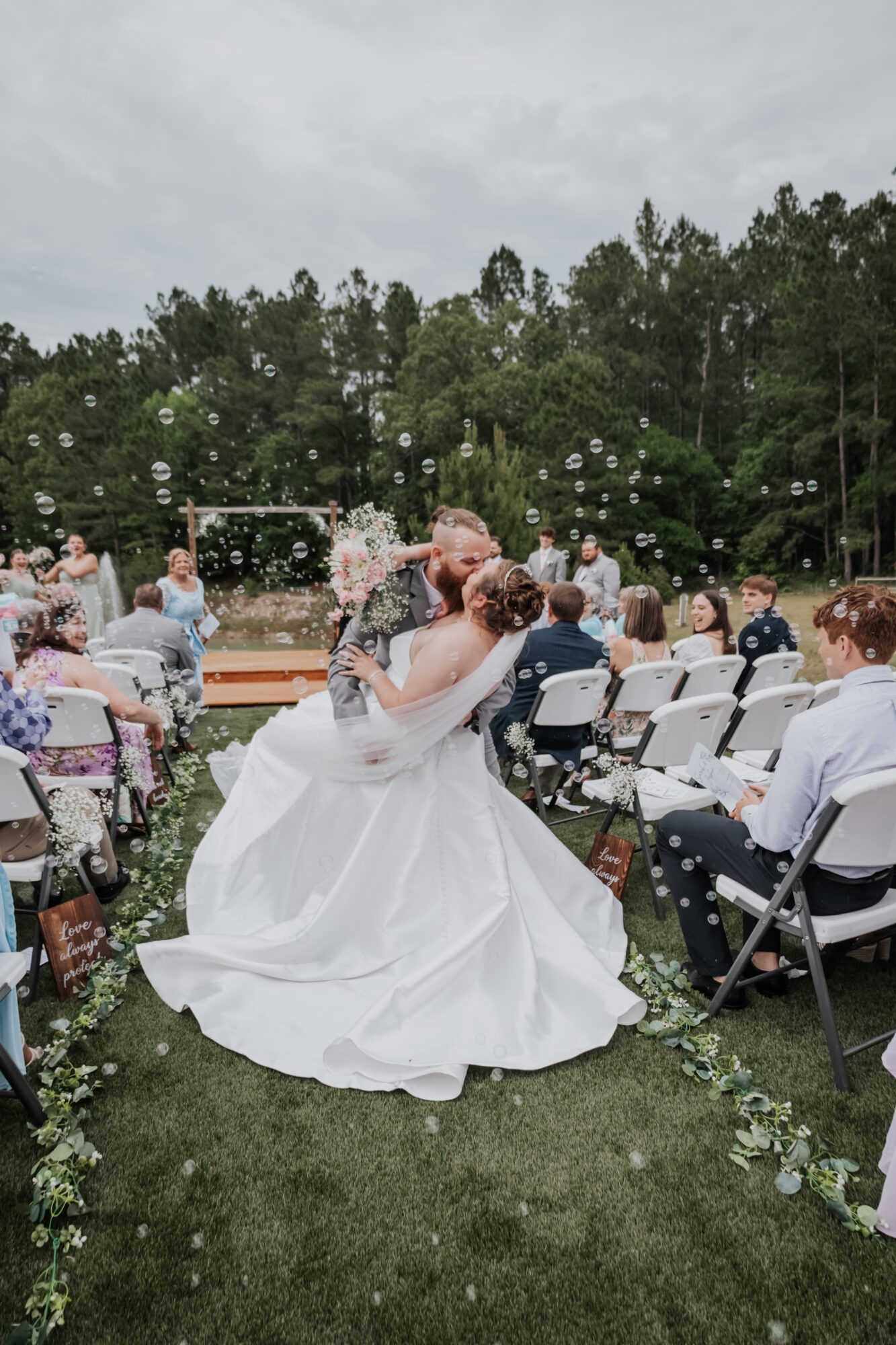 Bride and groom kiss outdoors during wedding ceremony with seated guests and trees in background.