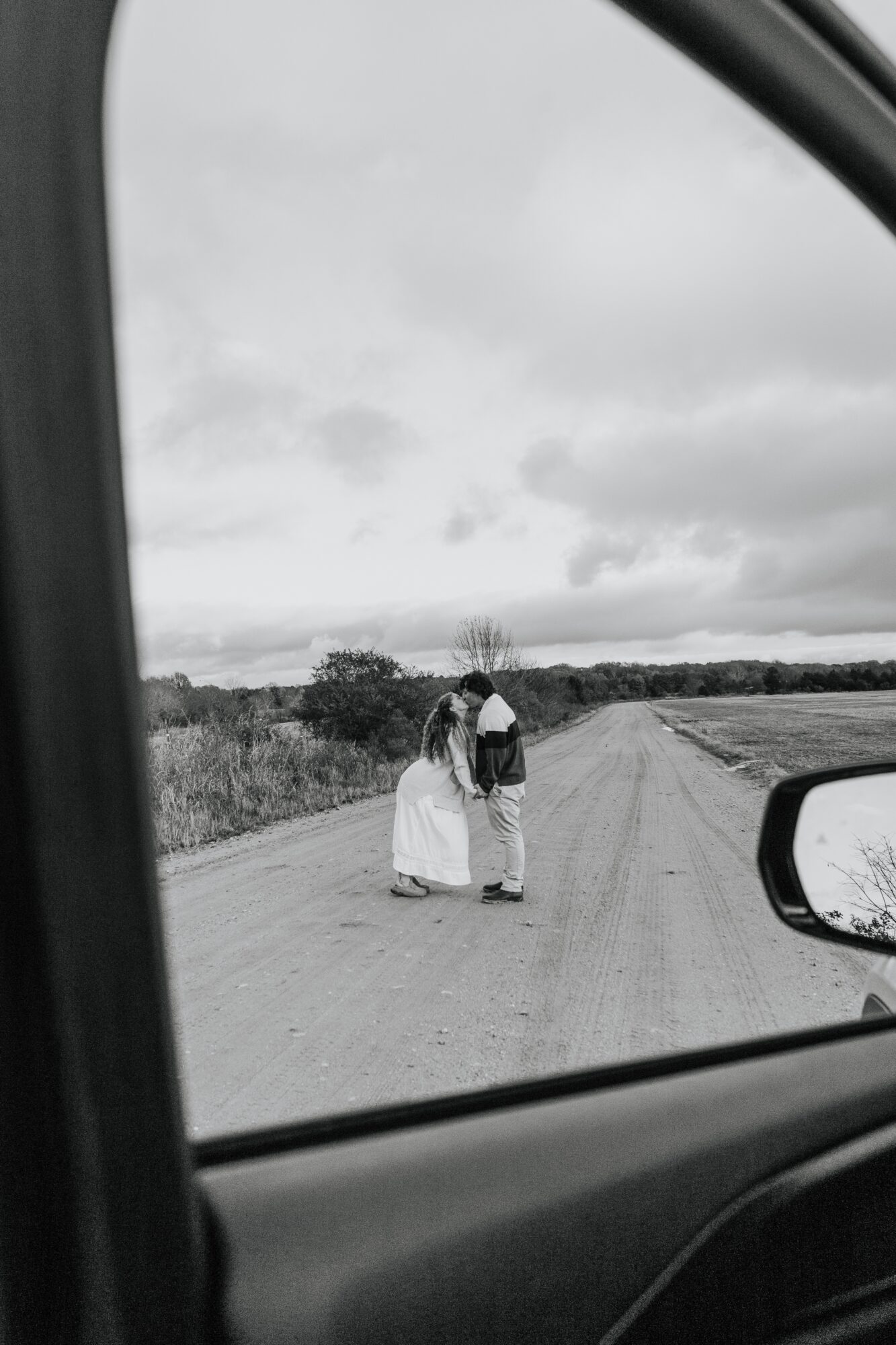 Two people walk on a dirt road with cloudy sky, seen from inside a vehicle, side mirror visible.