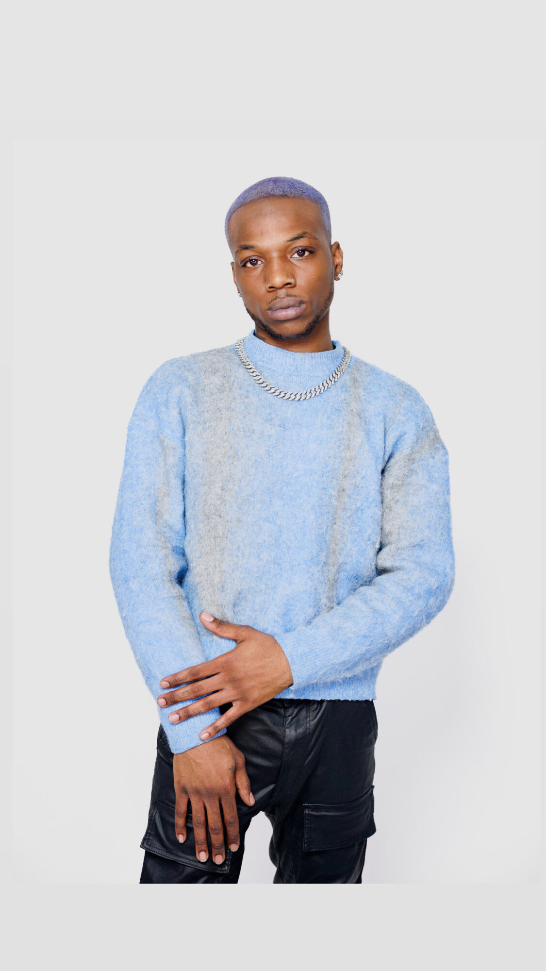 Person with short hair wearing a light blue sweater and black pants, standing against a plain background.