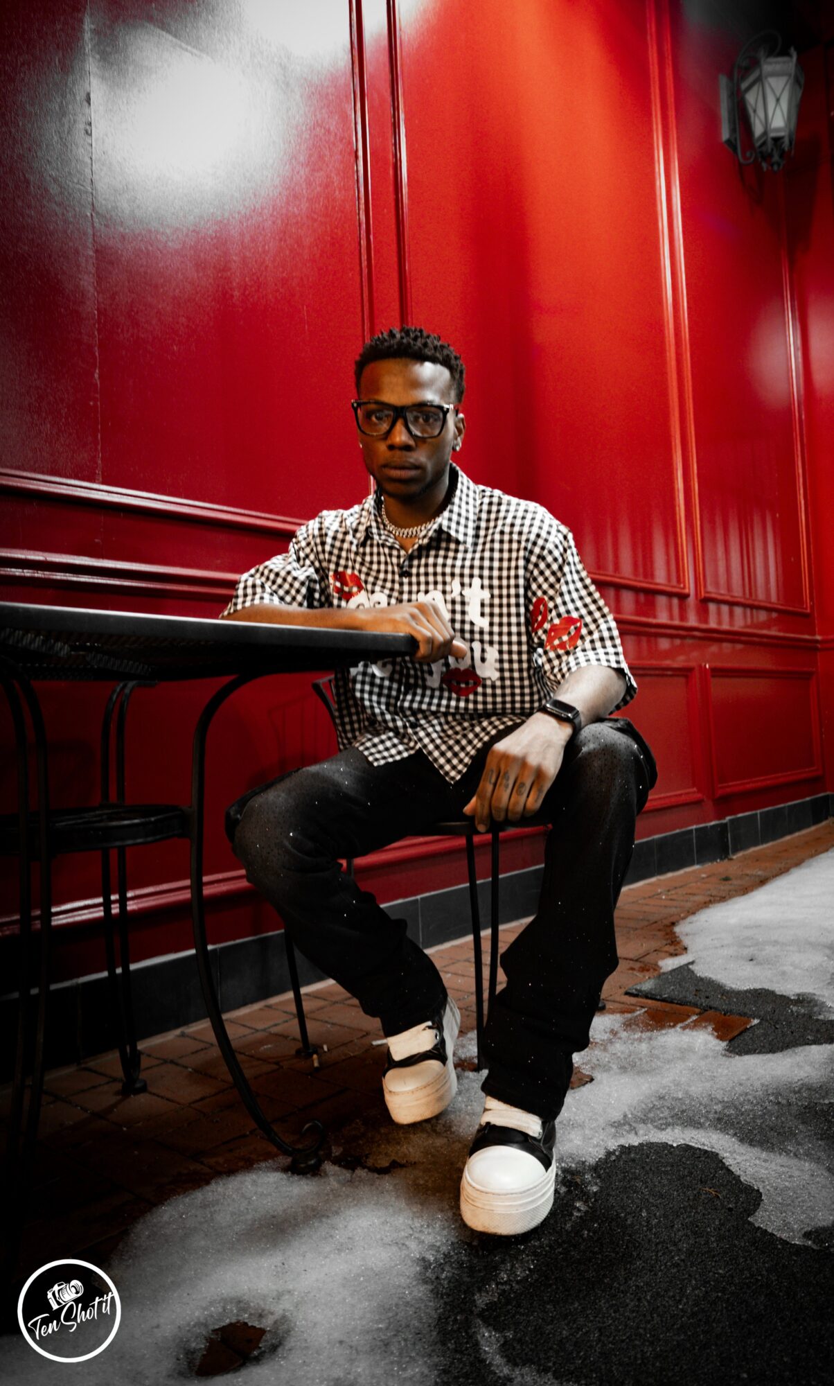 Person sitting on a chair against a red wall, wearing glasses, checkered shirt, and white sneakers.