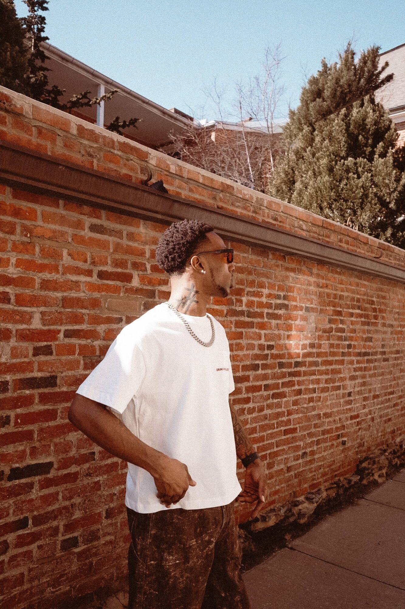 Young man with curly hair wearing a white t-shirt and patterned pants standing against a brick wall outdoors.