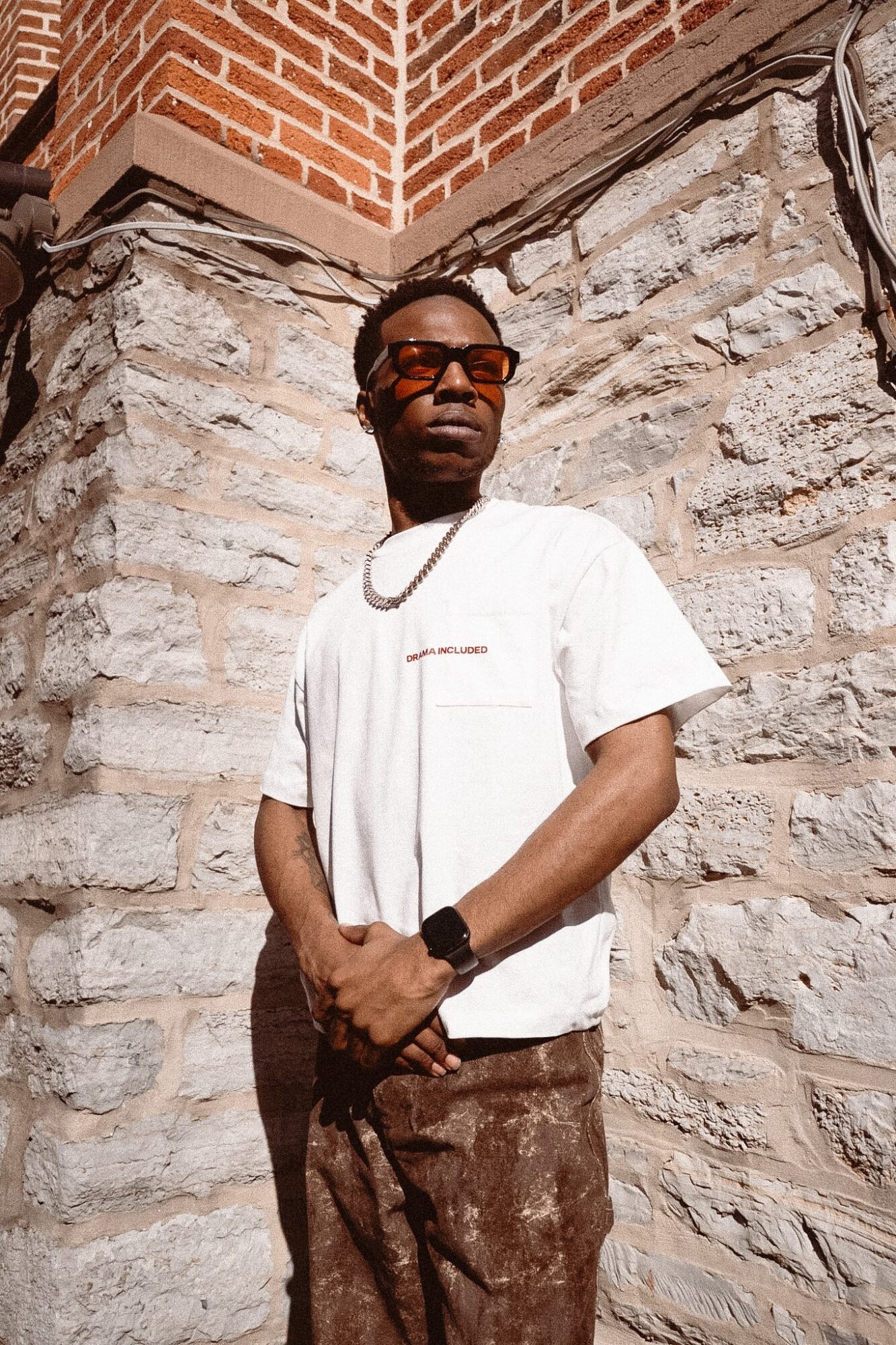 Man with sunglasses and a white t-shirt stands against a stone wall, looking to the side, sunlight on face.