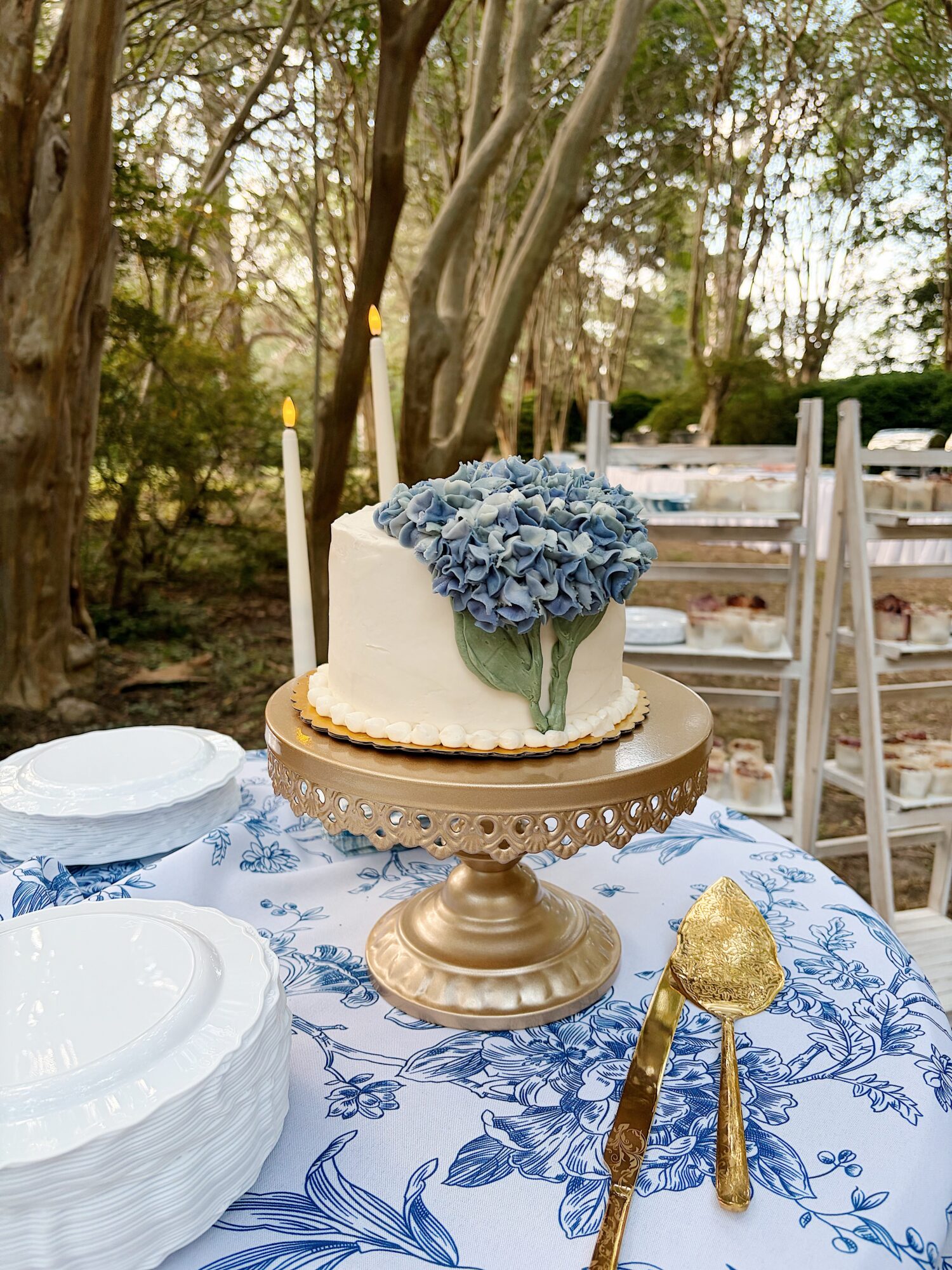 White cake decorated with blue flowers on a gold cake stand, surrounded by white plates and a gold spoon, outdoors.