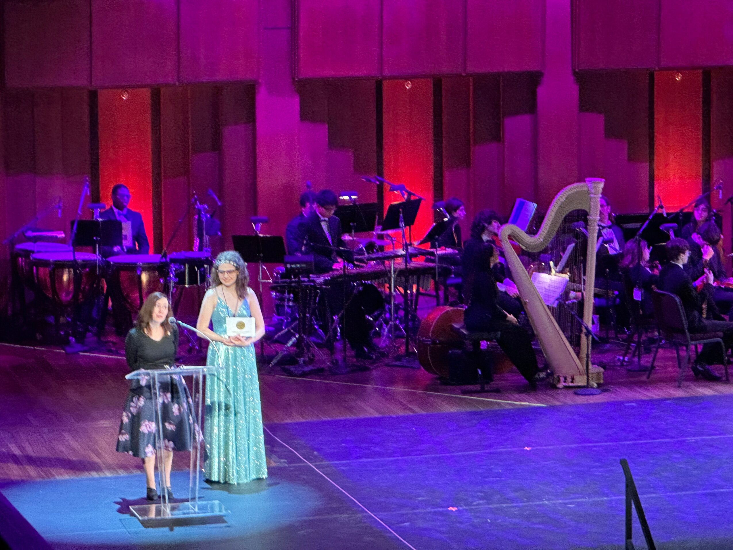 Two women stand on stage with musical instruments and a large harp behind them, in a concert hall with purple lighting.