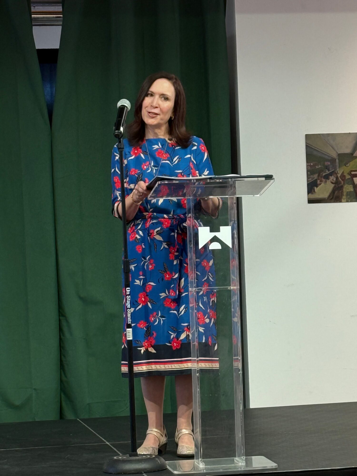 Woman in a blue floral dress stands behind a clear podium, speaking into a microphone, with a green curtain backdrop.