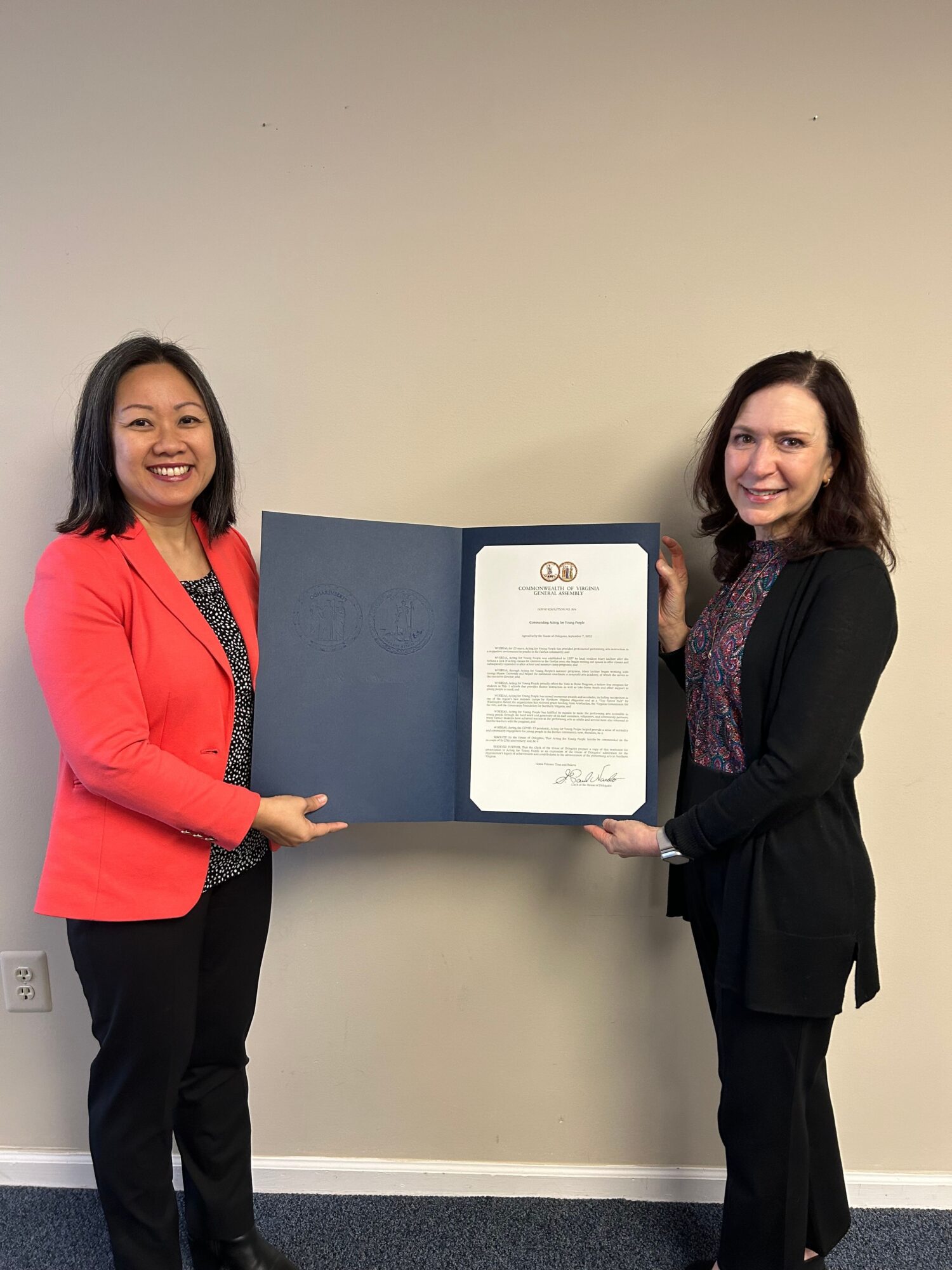 Two women standing indoors, one holding an open certificate or document, smiling at the camera.