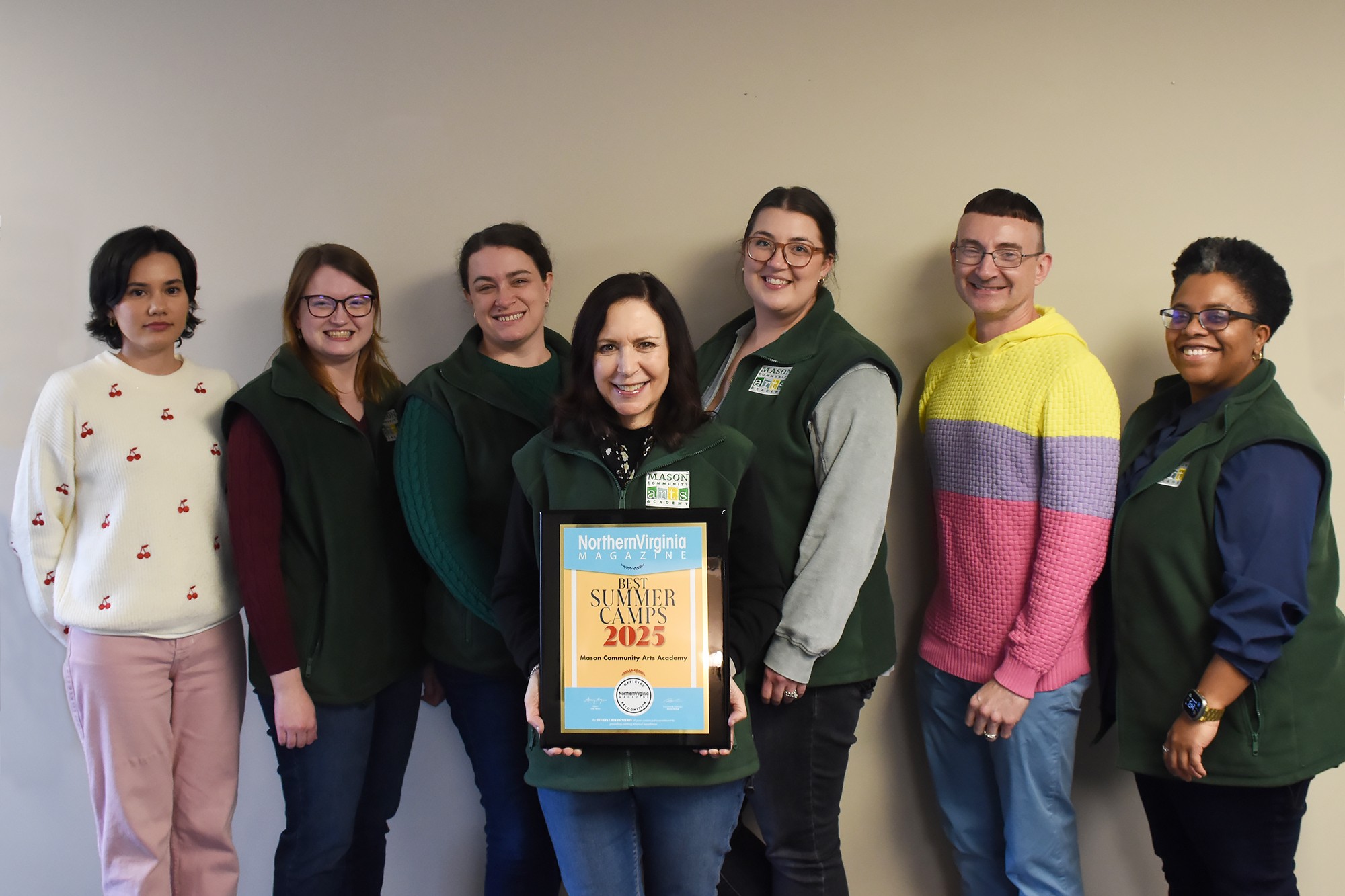 Group of seven people standing indoors, one holding a framed certificate, smiling, against a plain wall.