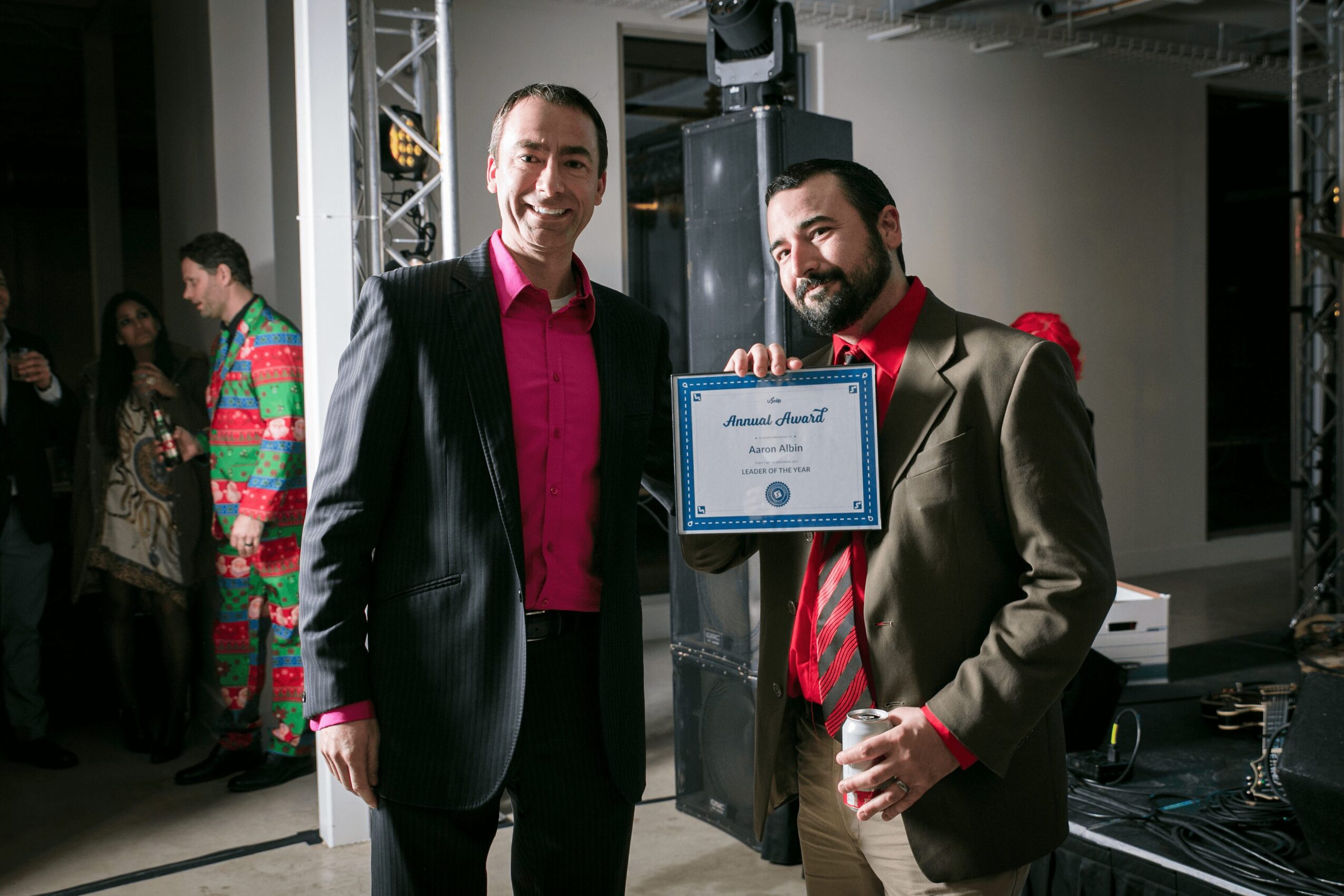 Two men in formal attire, one holding a certificate, smiling at an indoor event with other people in the background.
