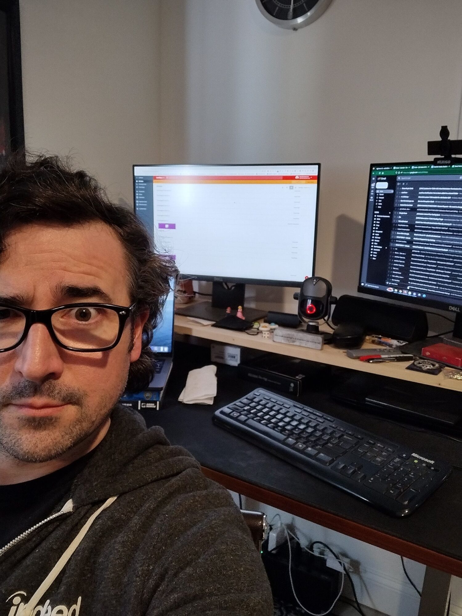 Person with glasses and dark hair at a desk with multiple monitors, keyboard, and computer equipment.