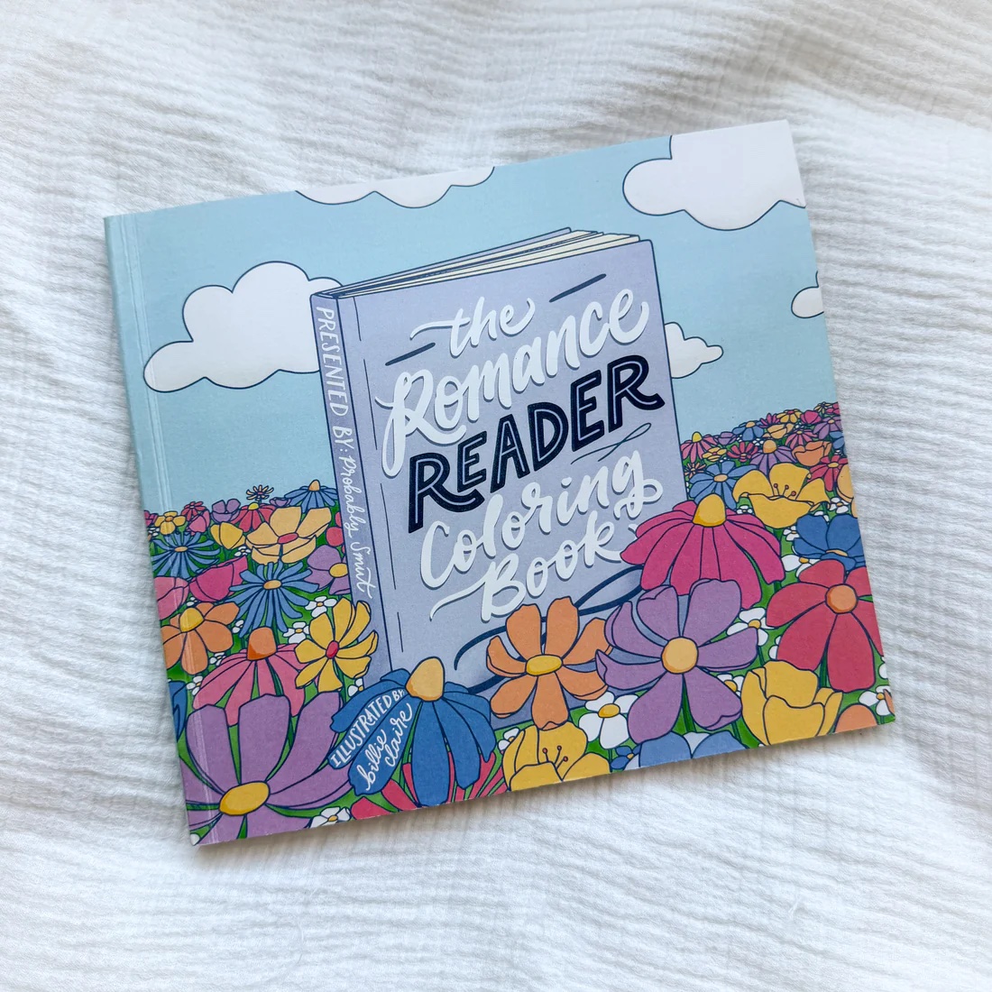 Coloring book titled 'The Romance Reader Coloring Book' with a sky and clouds background, surrounded by colorful flowers.