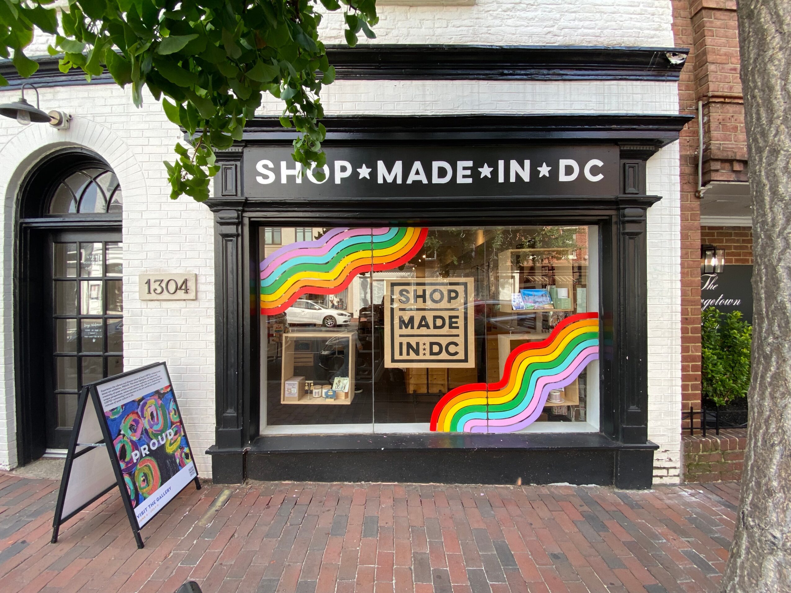 Storefront with sign reading 'SHOP MADE IN DC' and colorful rainbow decorations in window, brick sidewalk outside.