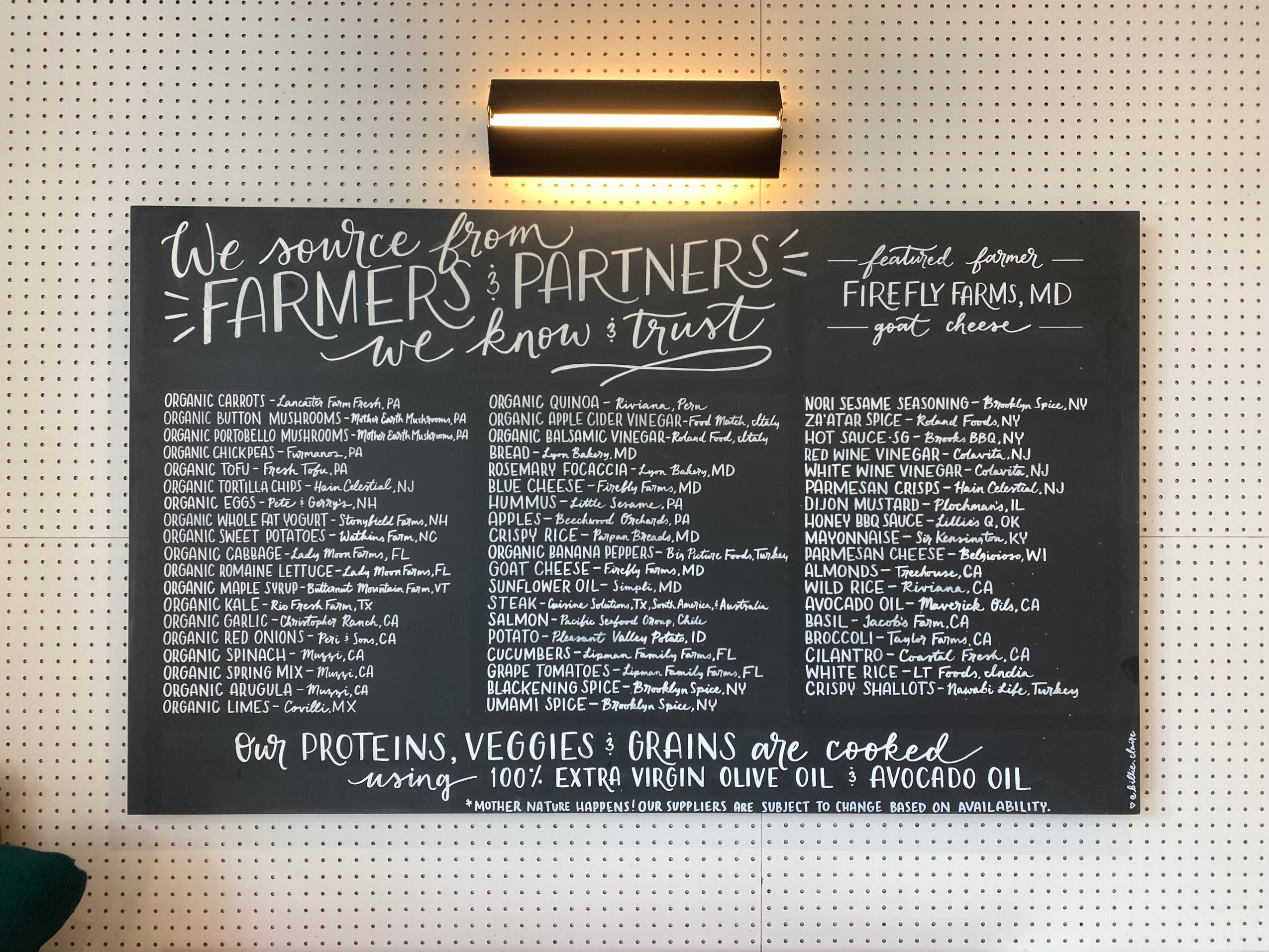 Blackboard menu listing farm products and partners, with handwritten text and decorative borders, illuminated by a light fixture above.