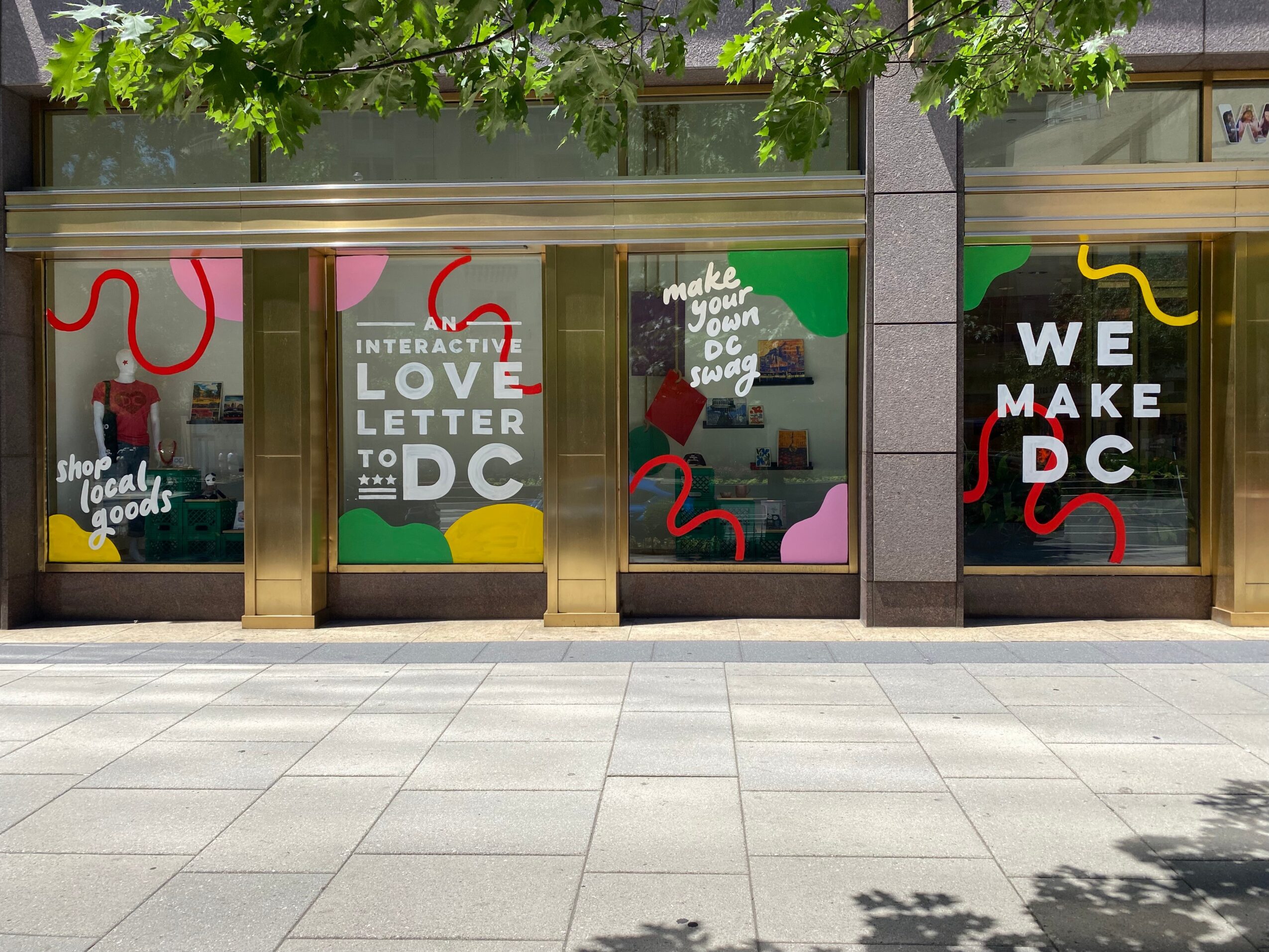 Storefront windows with colorful decorations and signs, trees above, and a sidewalk in front.