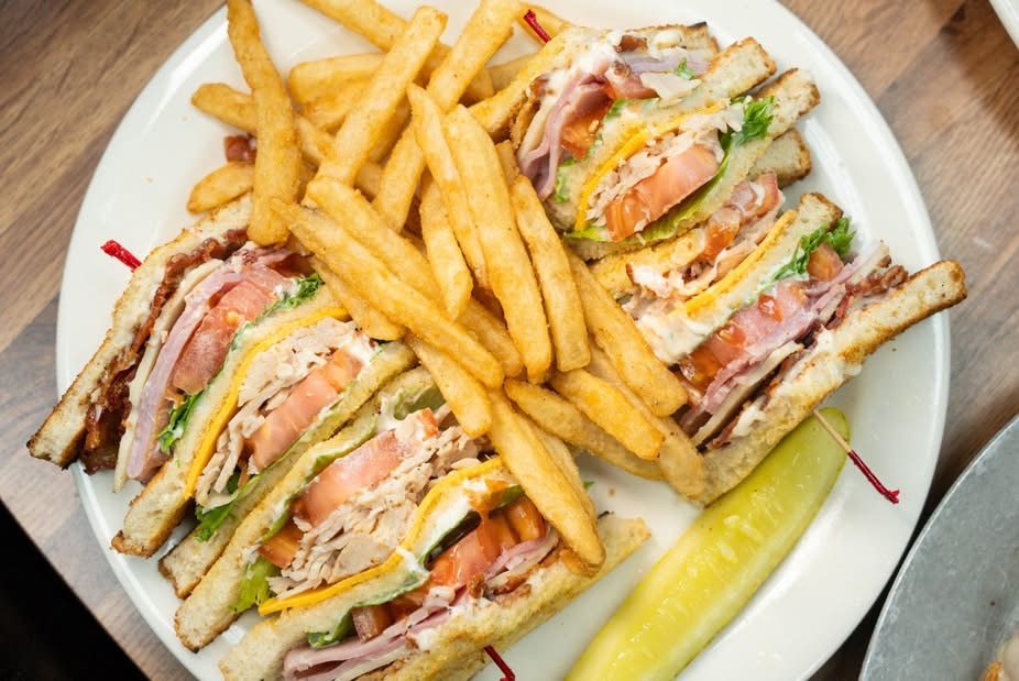 Club sandwich with fries and pickle on a white plate, viewed from above.