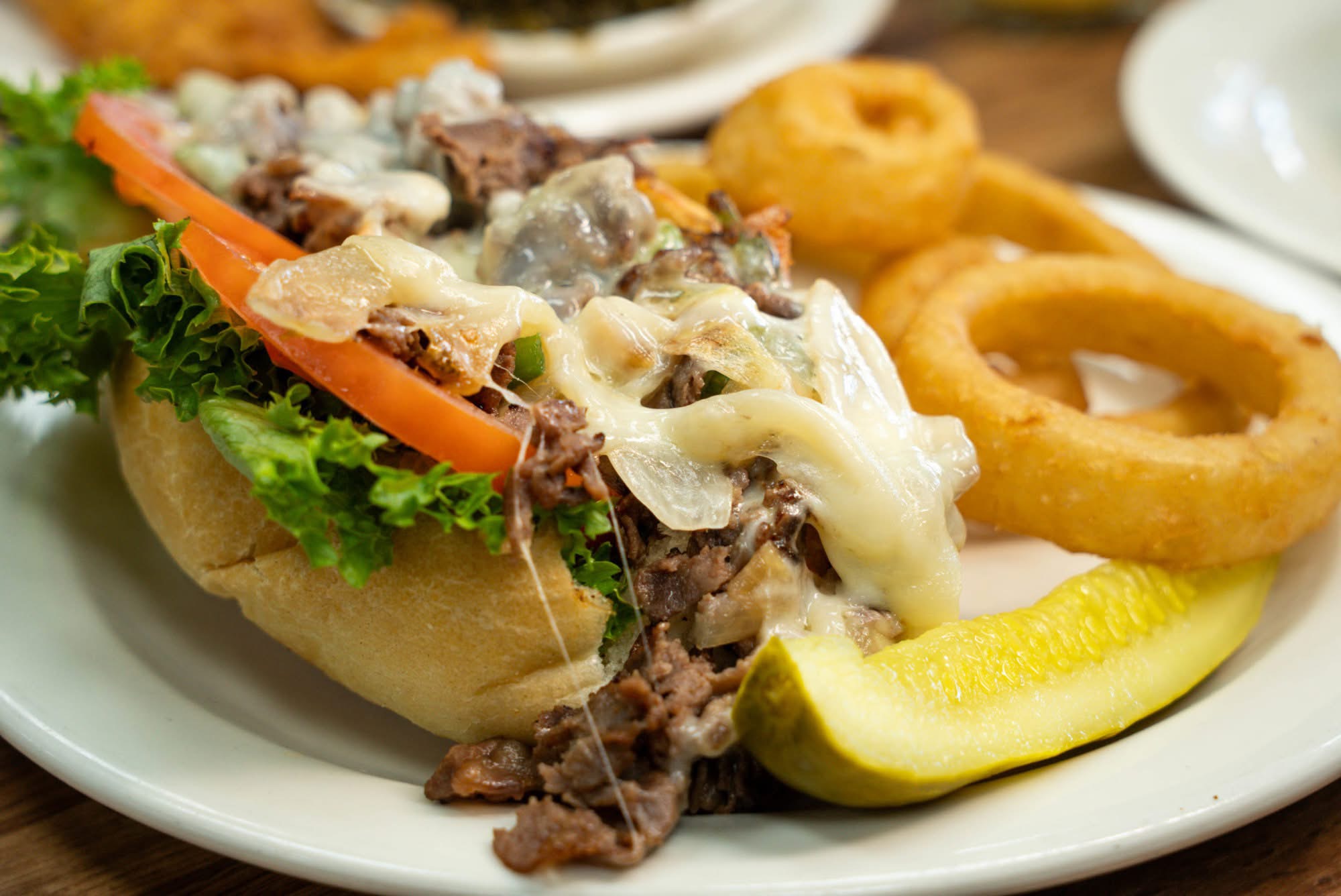 Close-up of a taco with lettuce, tomato, cheese, and meat, served with onion rings and a pickle on a white plate.