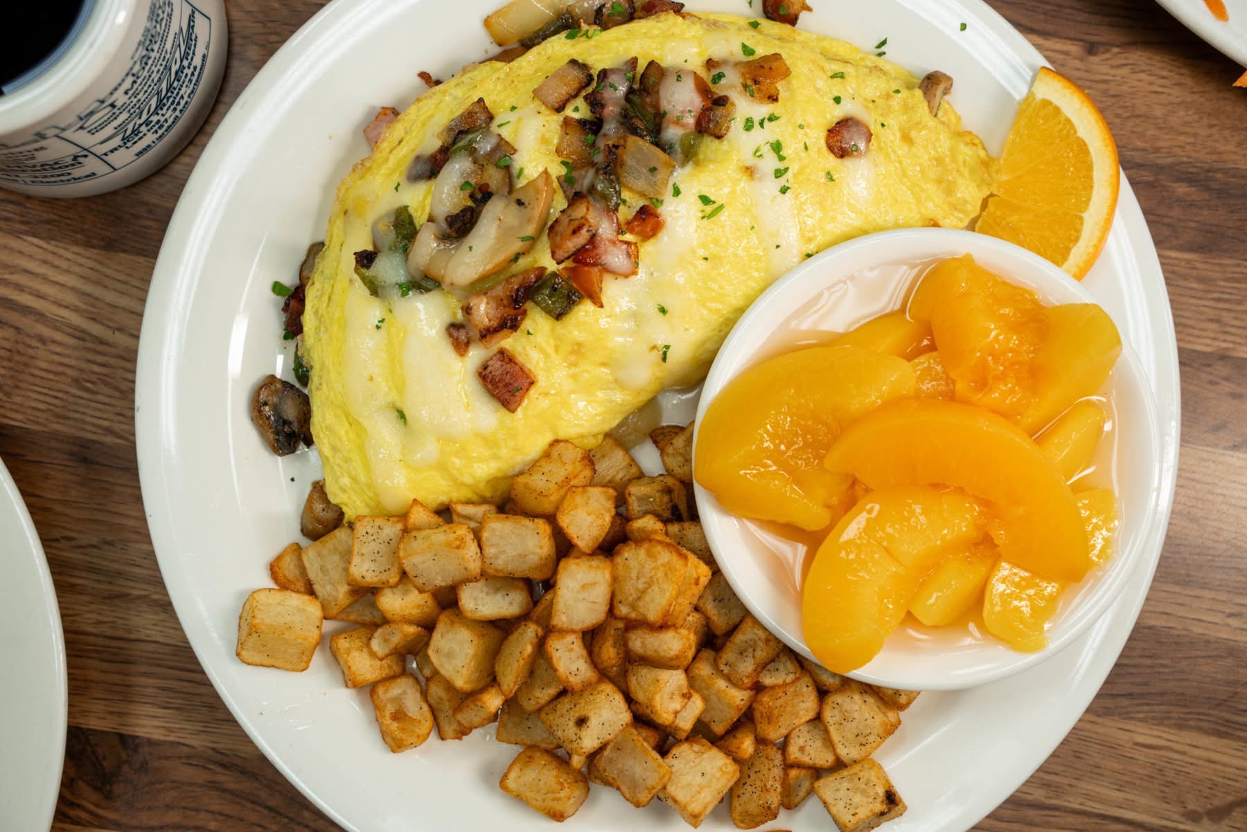 Omelette topped with vegetables and bacon, diced potatoes, and canned peaches in a bowl on a white plate, on a wooden table.