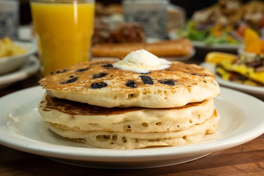 Stack of three pancakes with blueberries and butter on a white plate, breakfast table with orange juice and food in background.