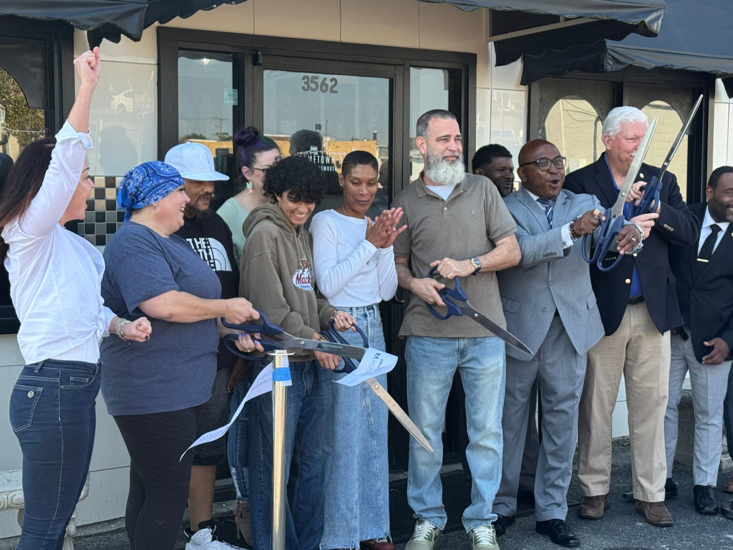 Group of diverse people gathered outside a building, some clapping and holding scissors for a ribbon-cutting ceremony.