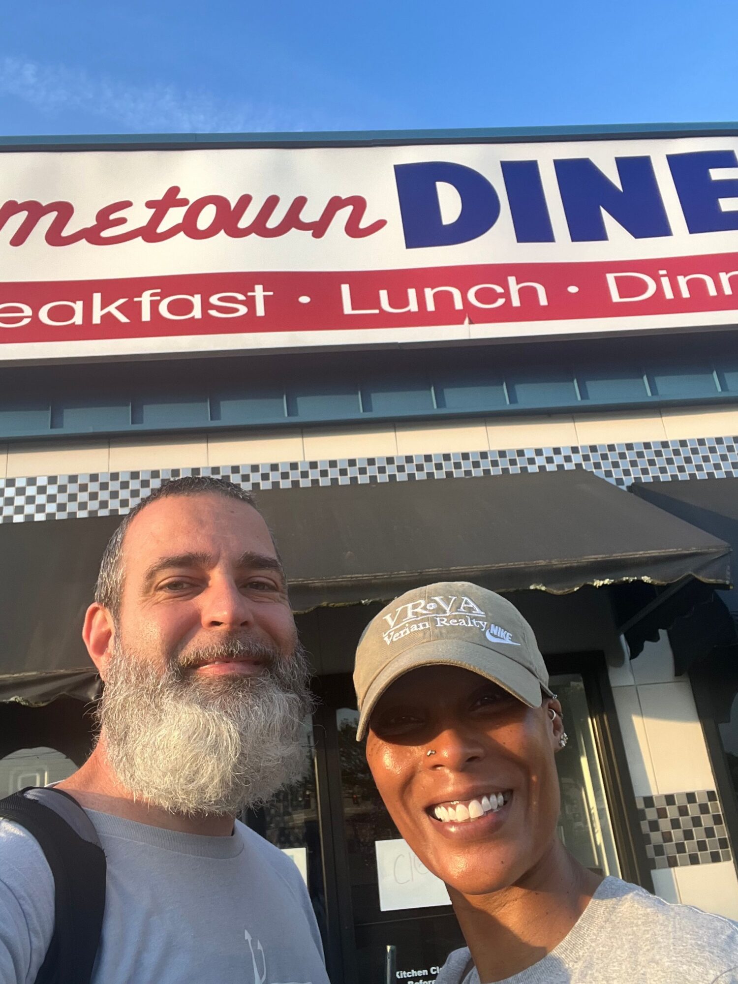 Two people smiling outside a restaurant with a sign reading 'Metown DINE' and menu items listed.