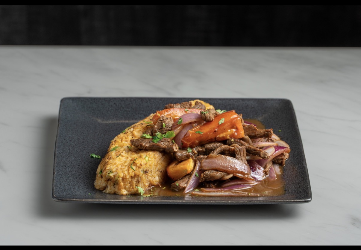 Plate with cooked meat, vegetables, and sauce on a black rectangular dish, on a light-colored surface.