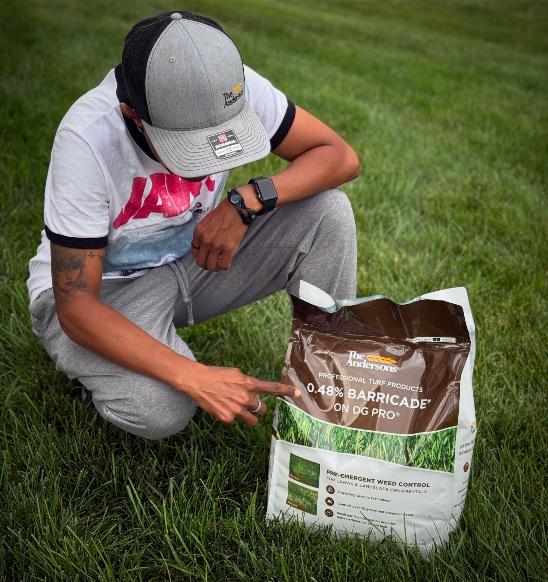 Person crouching on grass, wearing a cap, pointing at a bag of fertilizer or soil amendment, outdoors in a grassy area.