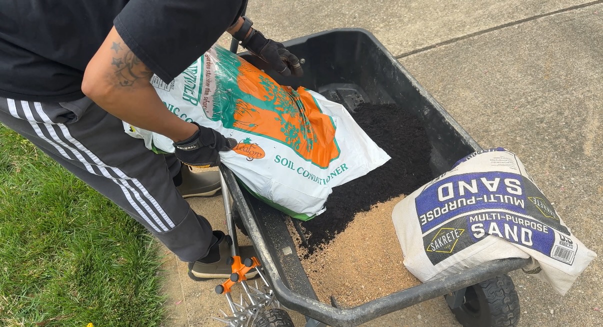 Person pouring sand from a bag into a wheelbarrow filled with sand and soil, outdoors on a sidewalk.