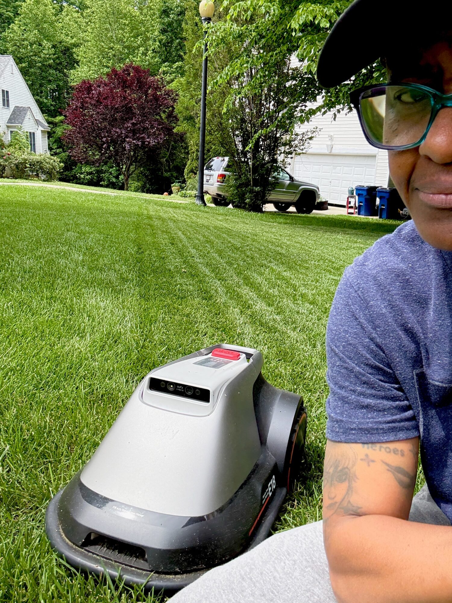 Person with glasses and tattooed arm operating a robotic lawn mower on a grassy yard, with trees and houses in background.