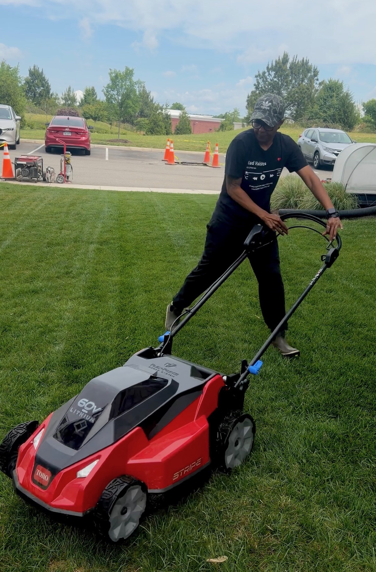 Person mowing grass with a red and black lawn mower on a green lawn, with parked cars and trees in background.