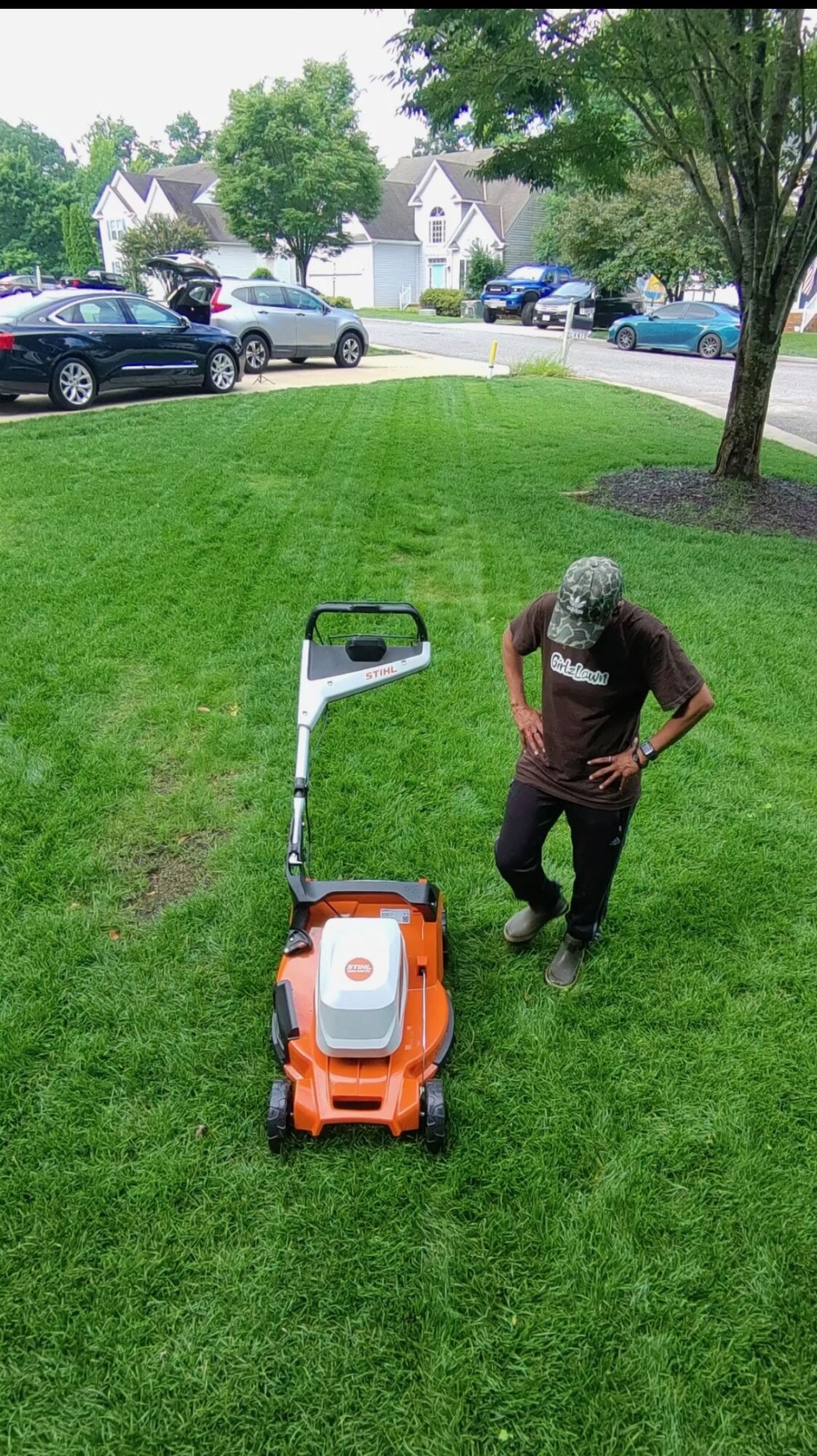Person standing on green lawn next to orange and white lawn mower, with trees and houses in background.