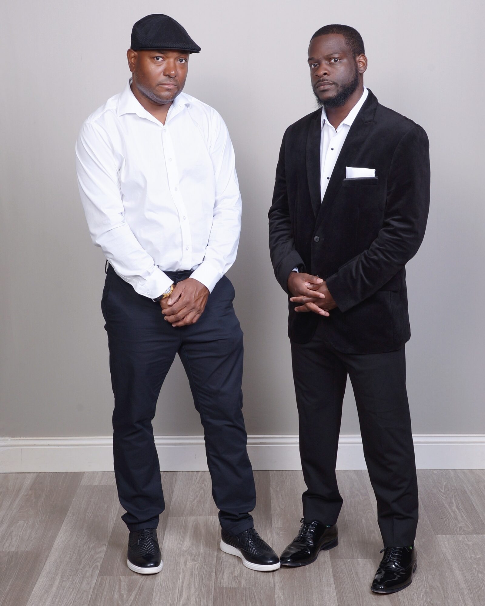 Two men stand side by side against a plain light gray background, dressed in formal attire, with hands clasped in front.