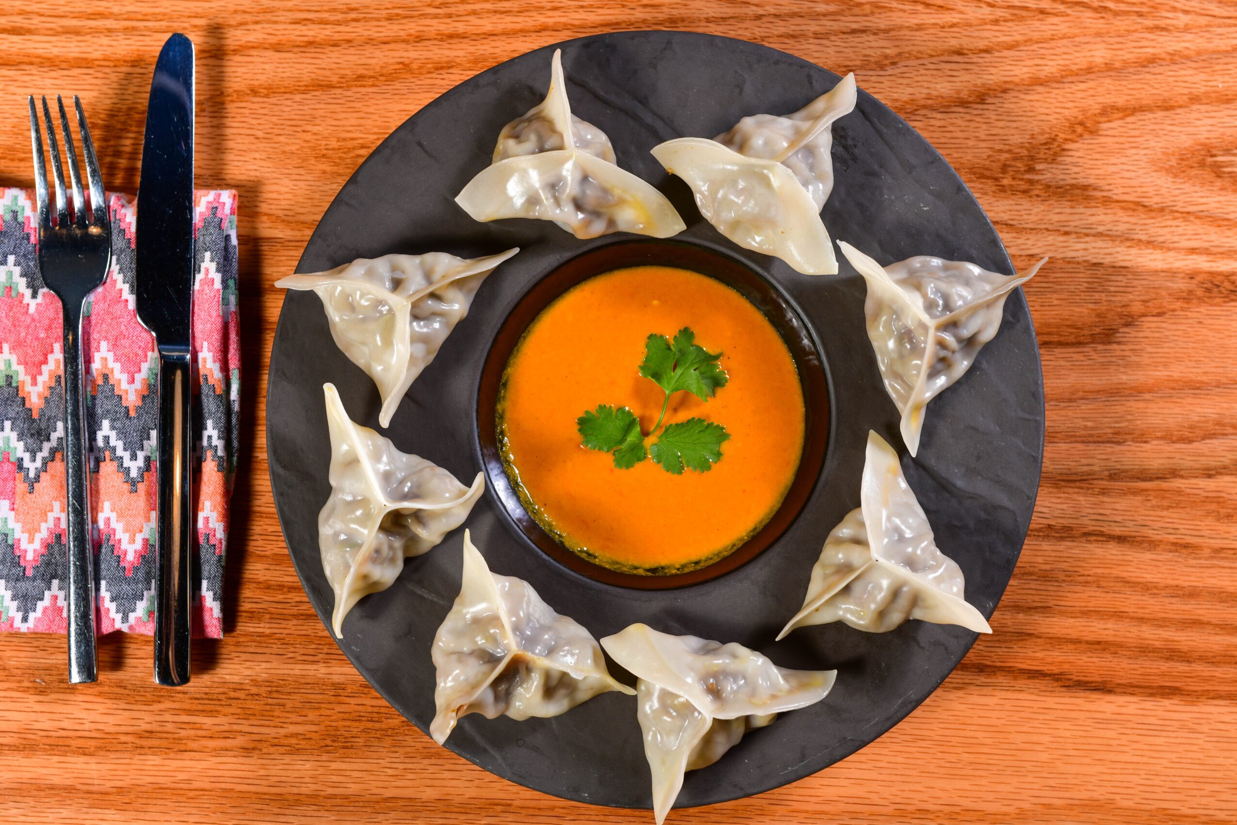 Plate with dumplings around a bowl of orange soup garnished with cilantro, on a wooden table, with utensils on a patterned napkin.