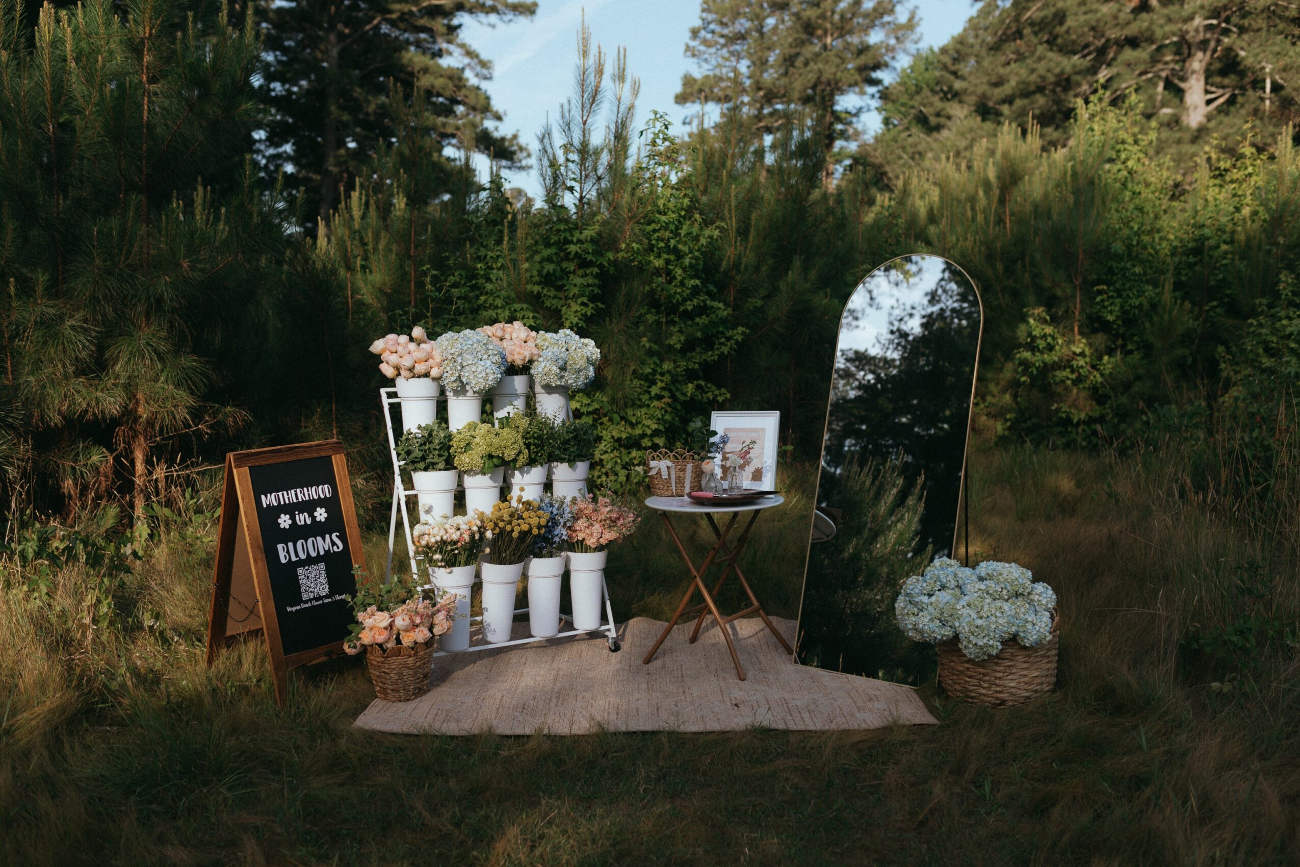 Outdoor floral display with white flowers, a mirror, and a small table, set on grass with trees in background.