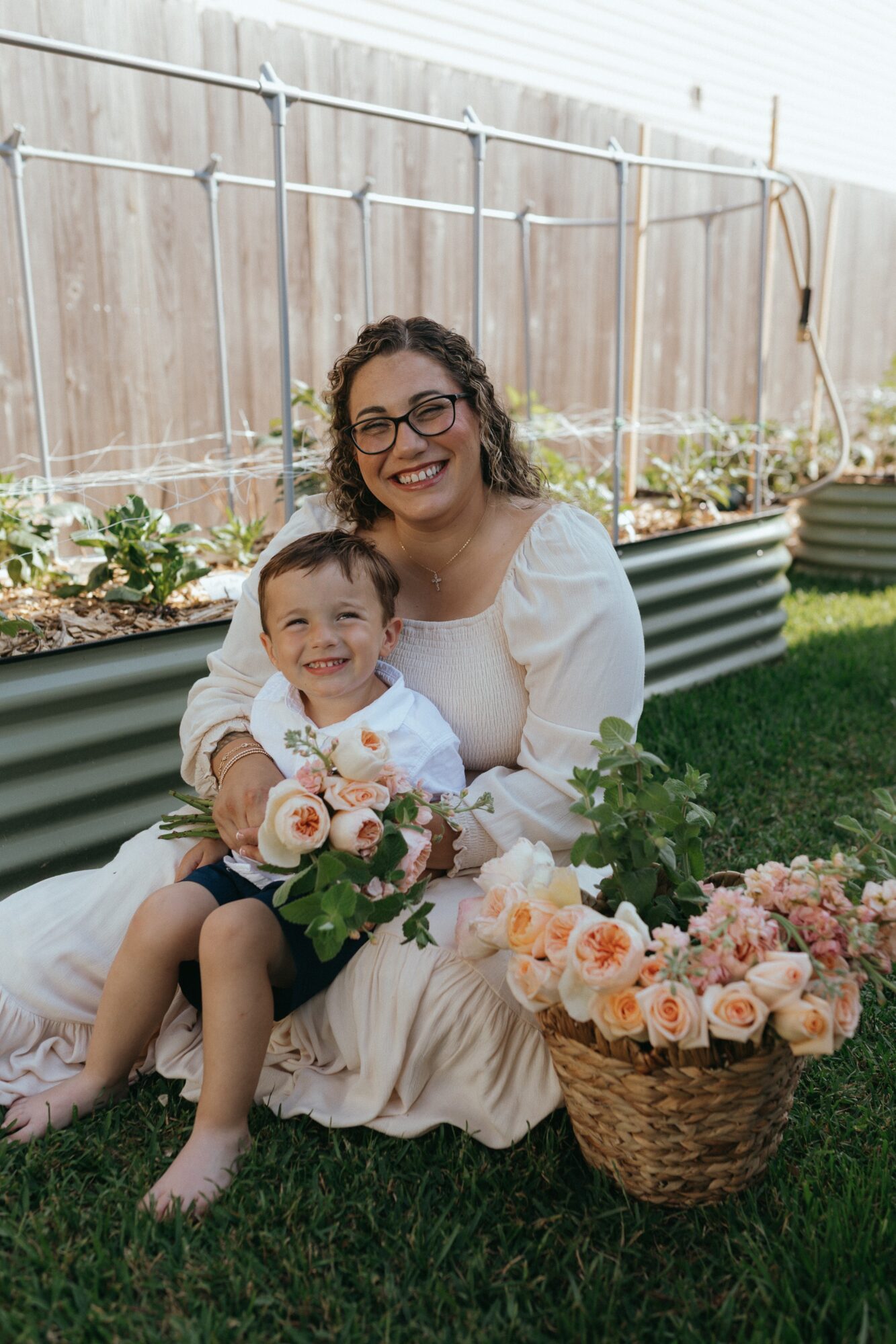 Woman and child sitting on grass with flowers and basket, smiling outdoors near a wooden fence.