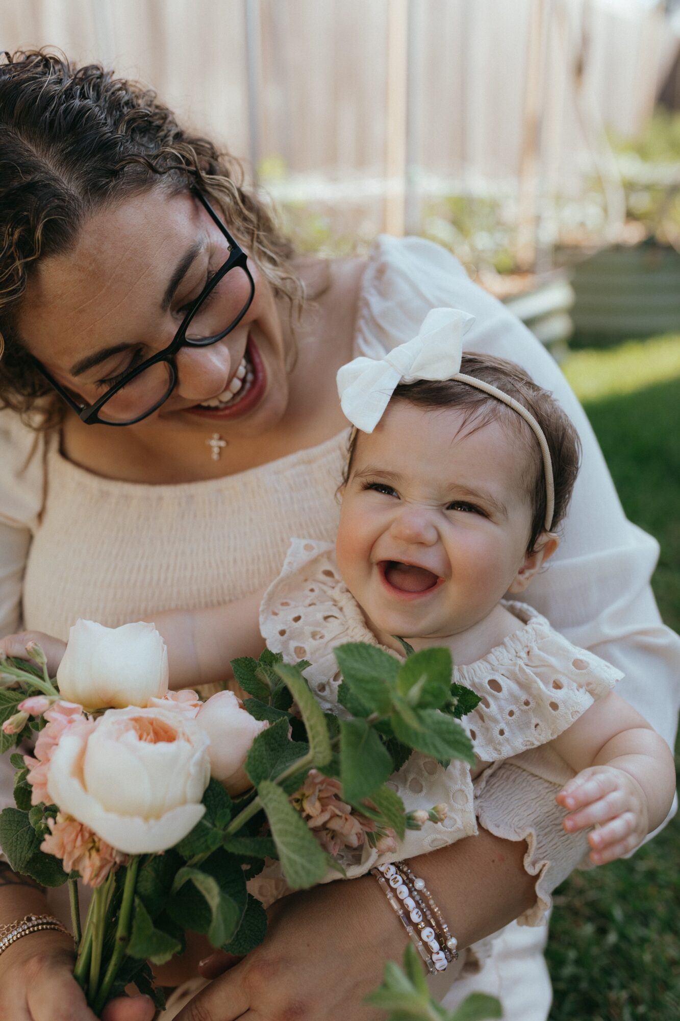 Woman holding smiling baby girl with flowers outdoors, woman wearing glasses and a white top, baby in a light dress with a bow.