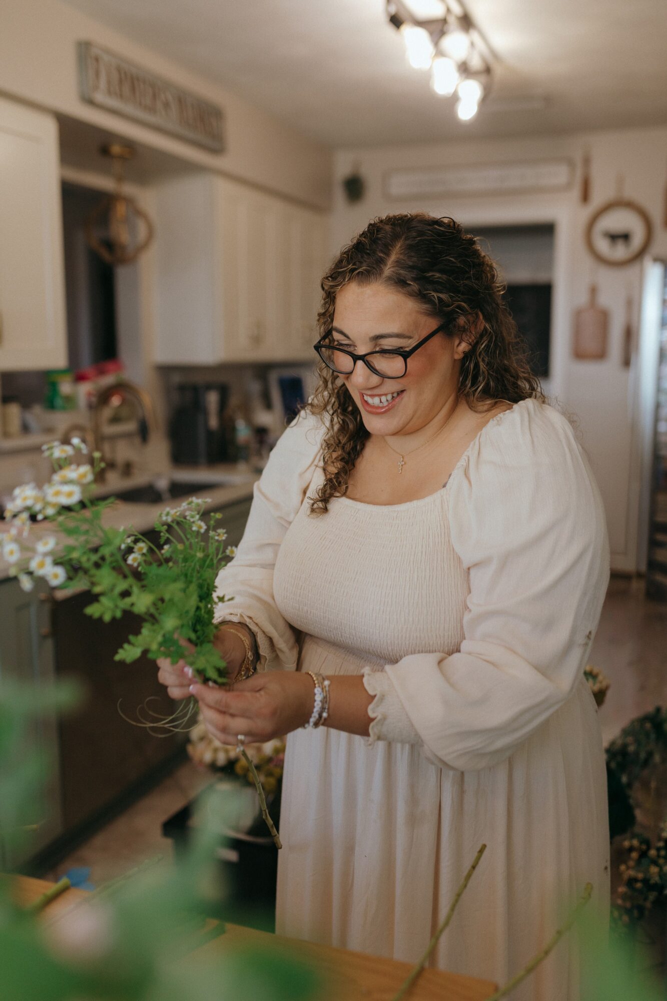 Woman with glasses smiling in a kitchen, holding a plant, wearing a white dress with puffed sleeves.
