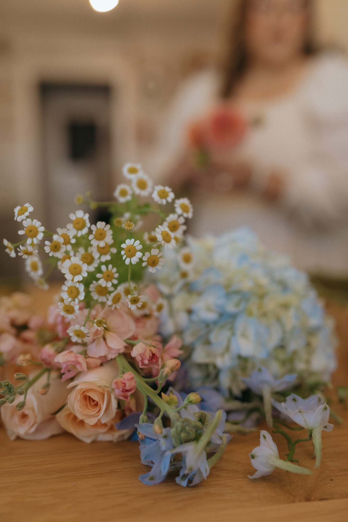 Bouquet of pink roses, white daisies, and blue hydrangeas on a wooden table with a blurred person in the background.