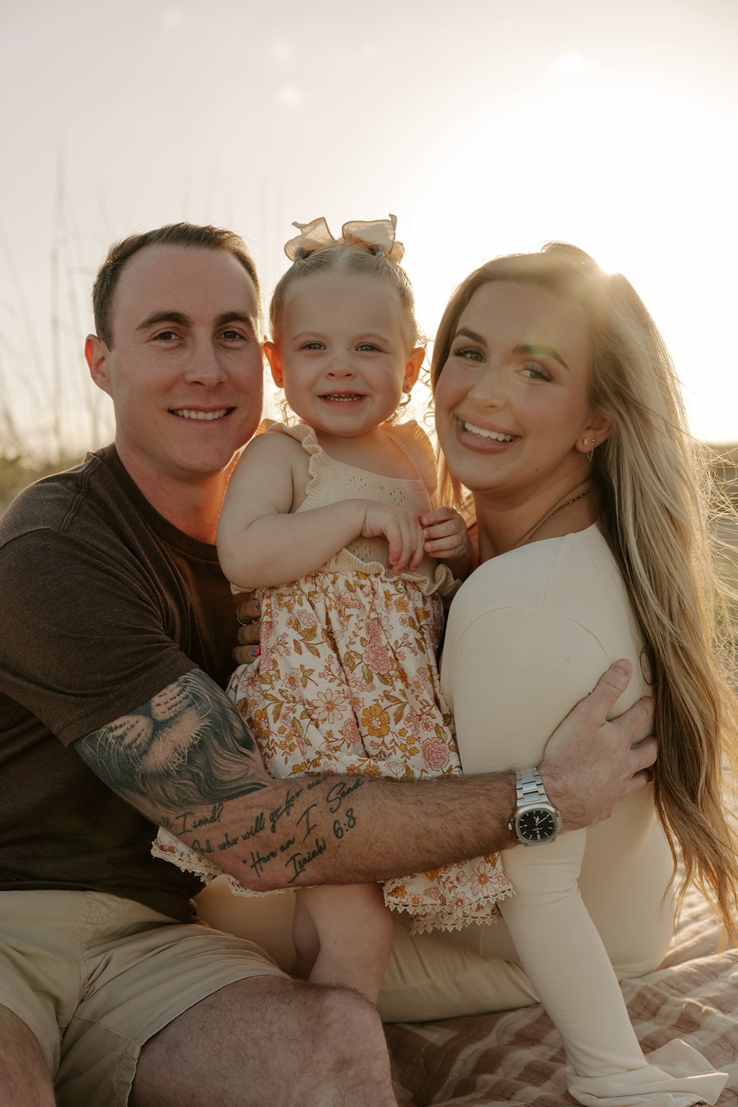 Family of three outdoors, smiling, with sunlight behind them, woman holding a young girl, man with tattoos, all close together.