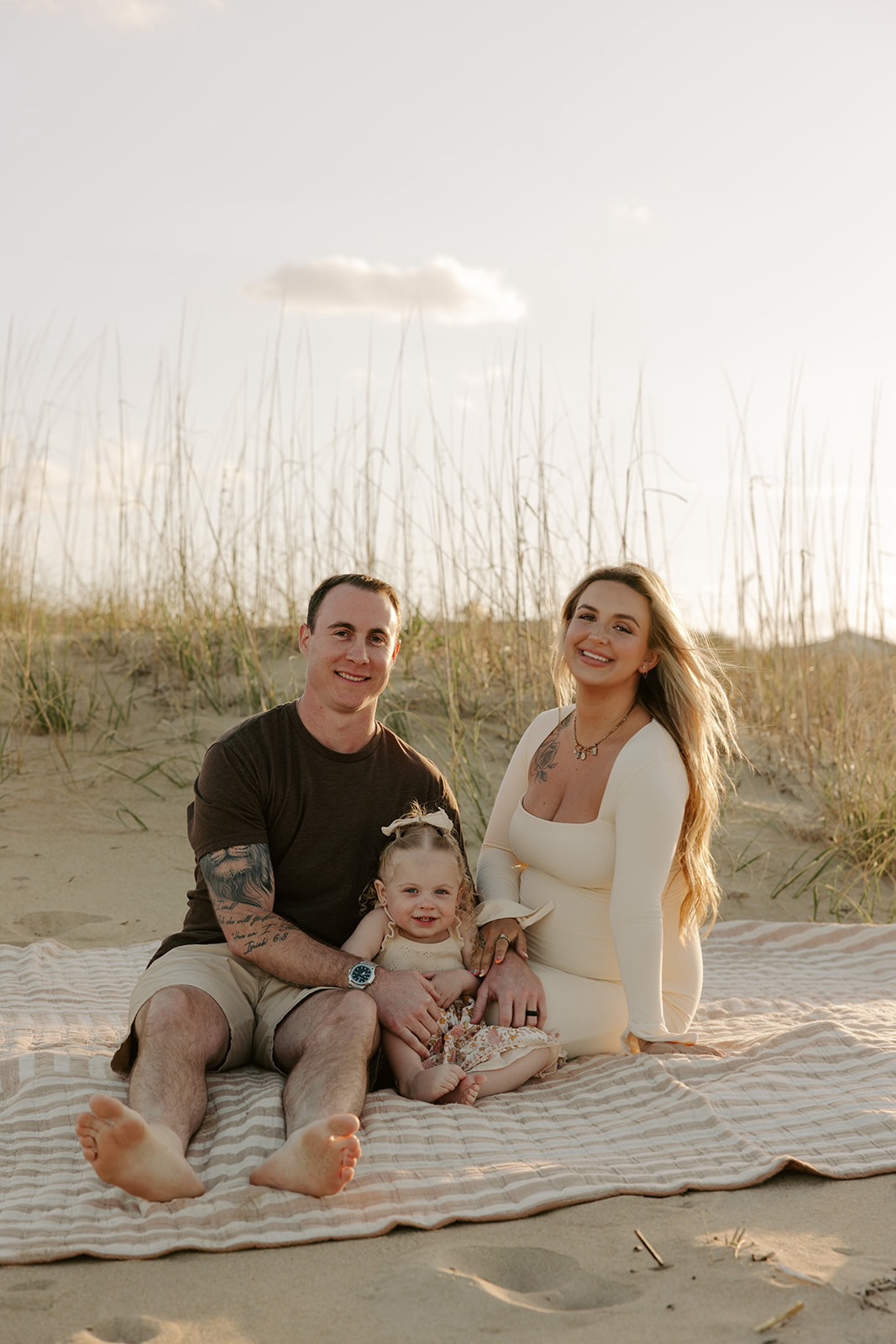 Family of three sitting on a blanket outdoors, smiling, with sand dunes and tall grass in the background.