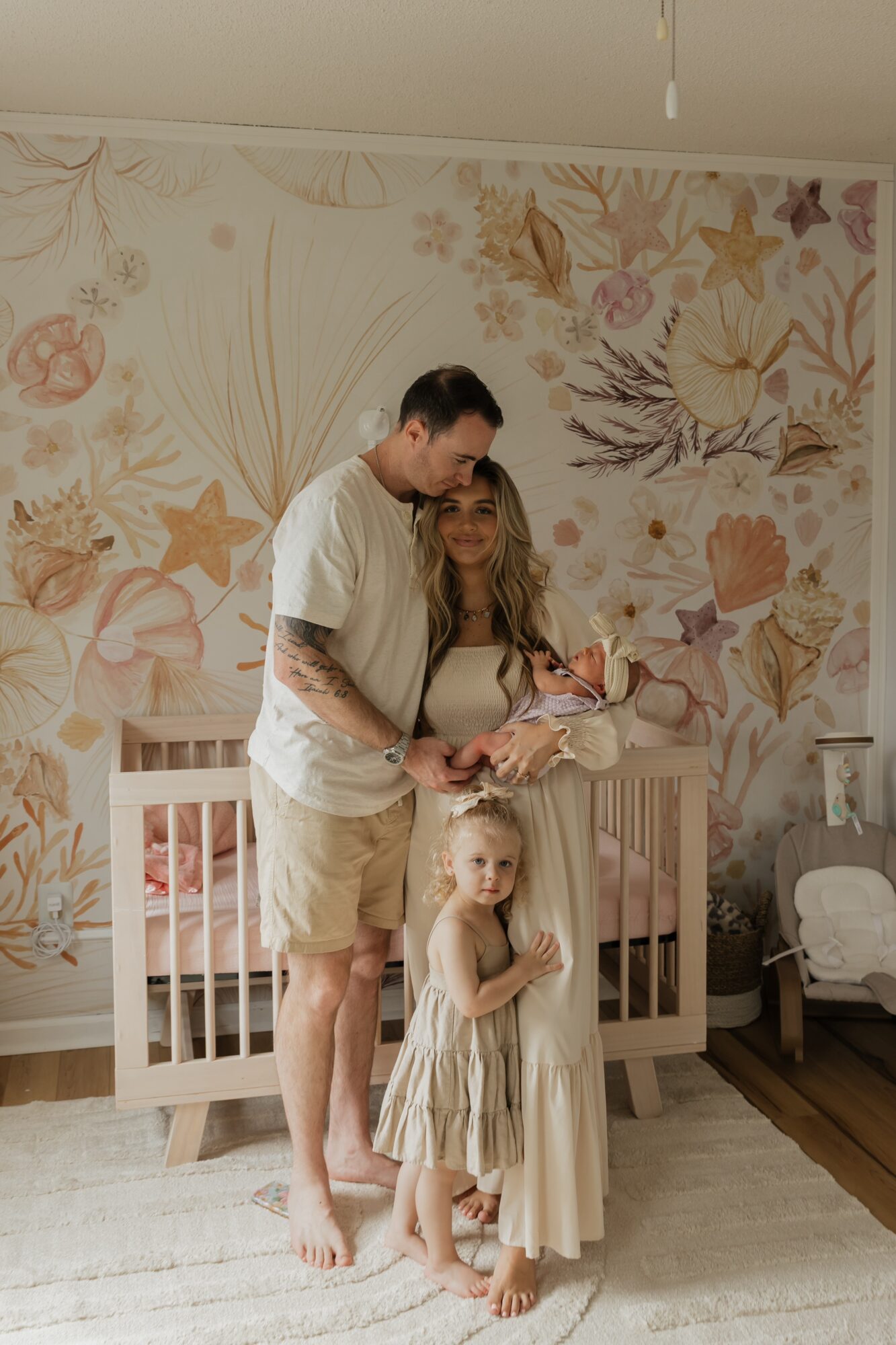 A man and two young girls stand in a bedroom with a floral wallpaper background. The man holds a book, and the girls stand nearby.
