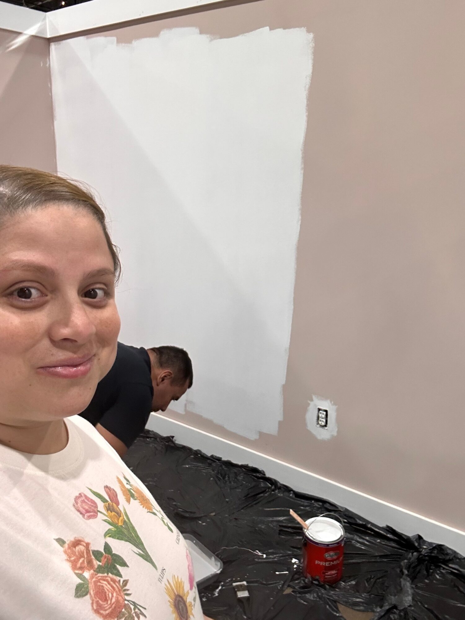 Person with short hair and floral shirt taking a selfie, another person working on wall in background, room under renovation.