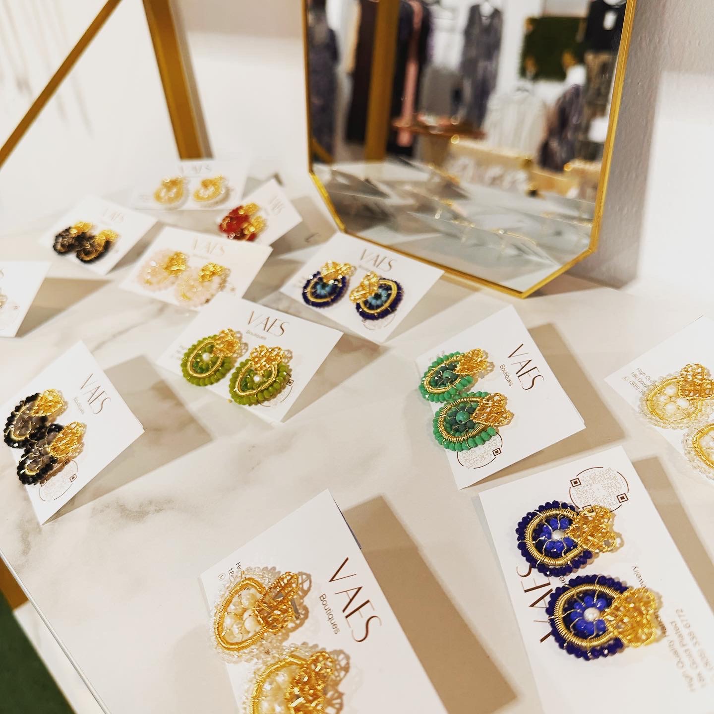 Display of colorful earrings on white cards, with a mirror reflecting a clothing store interior in the background.