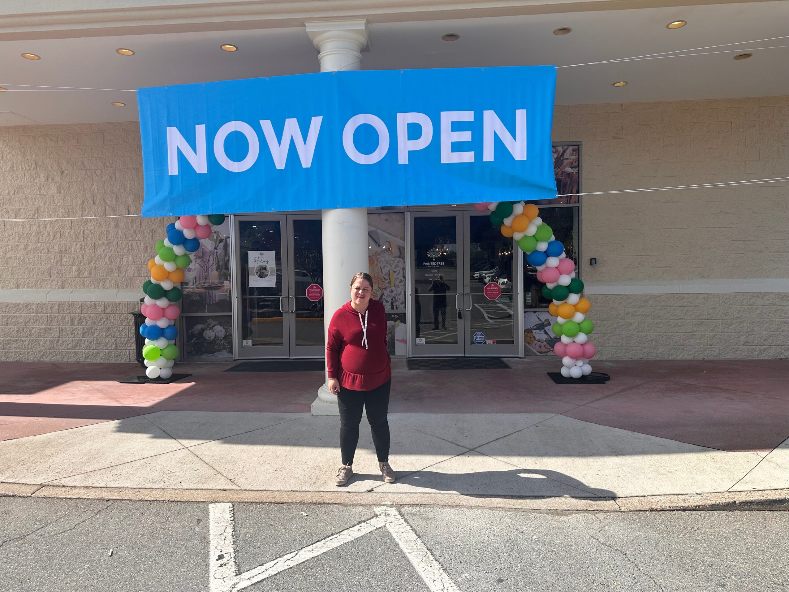 Person standing outside a store with a large blue banner reading 'NOW OPEN' and balloon decorations on either side.